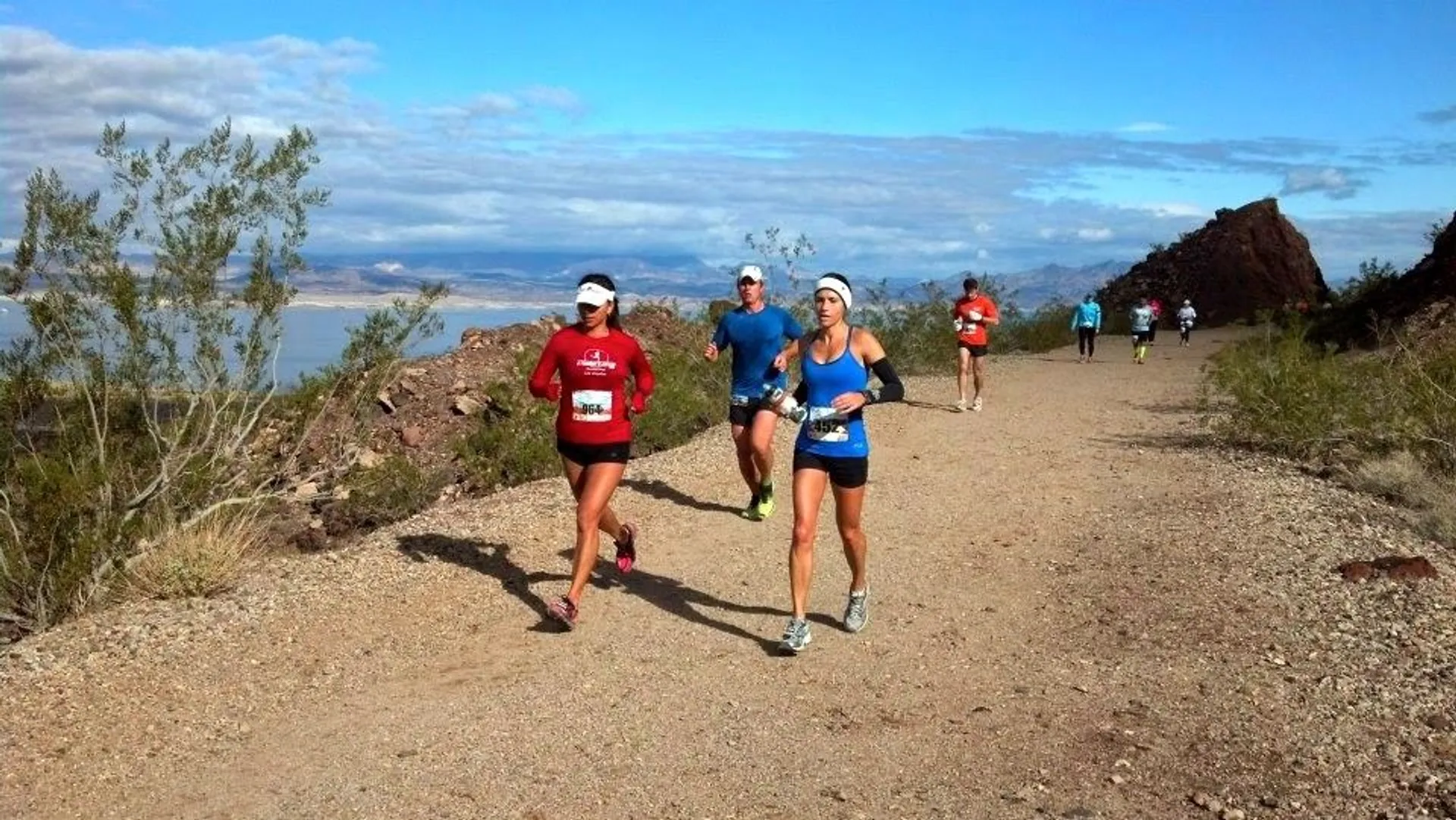 The image shows a group of people running on a dirt trail with a scenic background