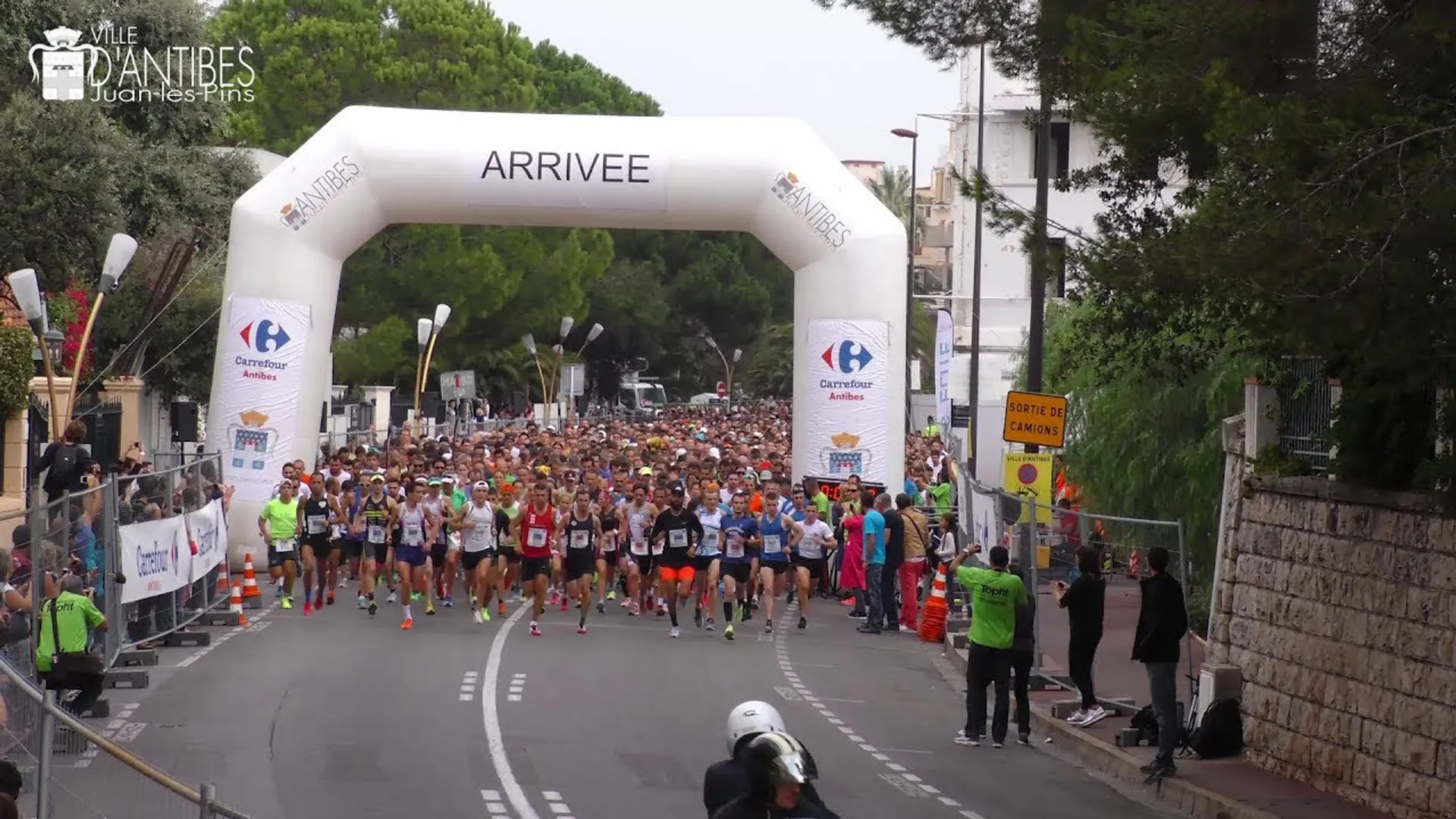 The image shows a road running event with a large group of runners at the starting