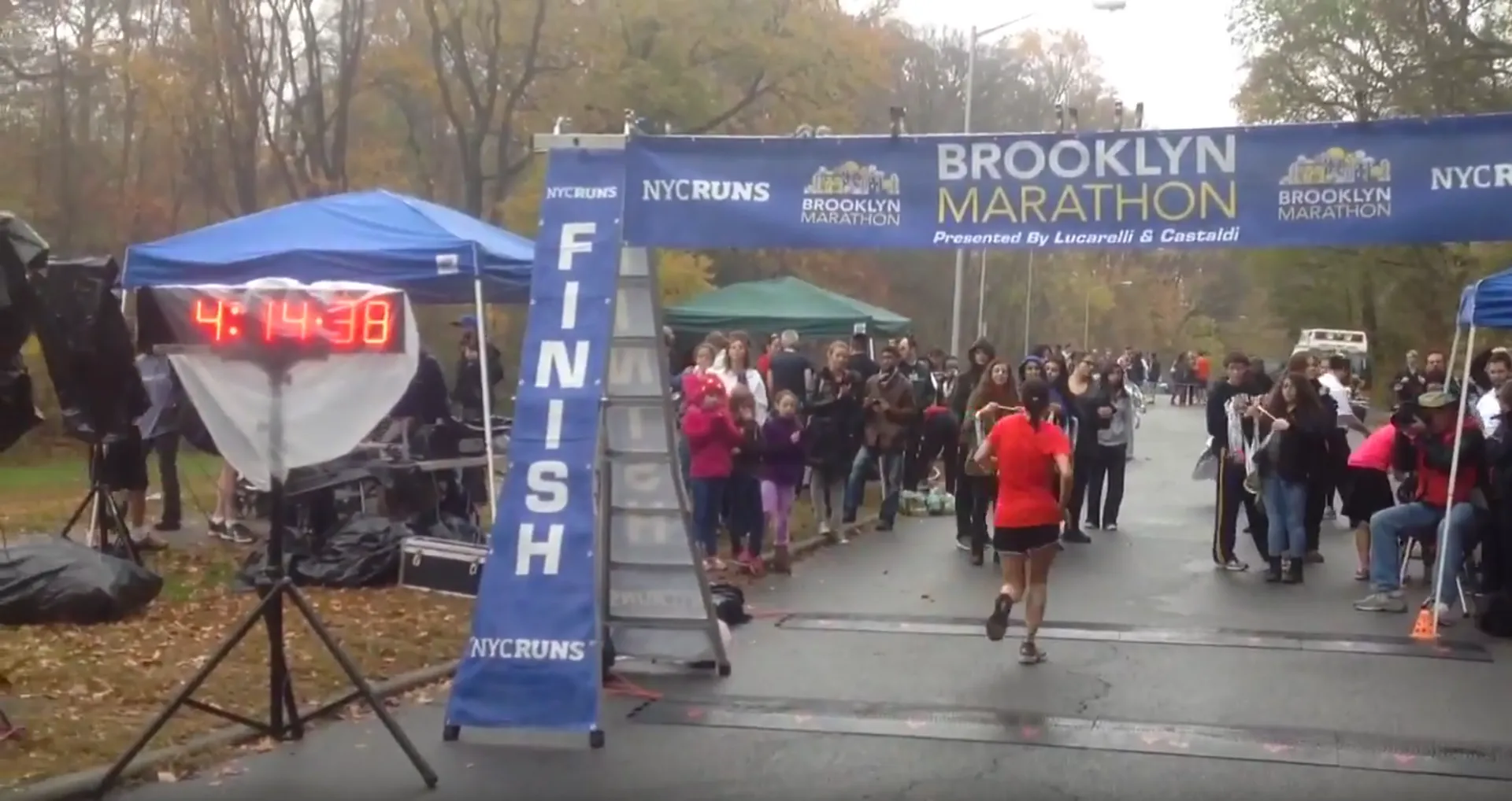 The image shows a marathon event, specifically the Brooklyn Marathon, with a runner crossing