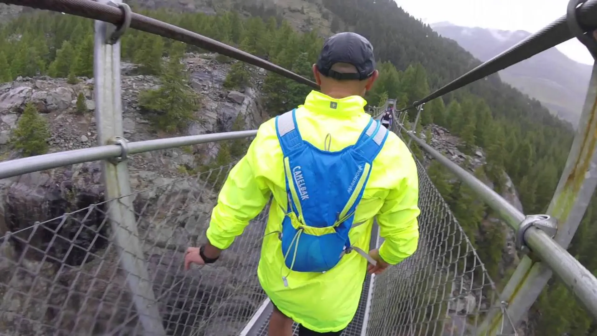The image shows a person from behind walking on a suspension bridge in a mountainous