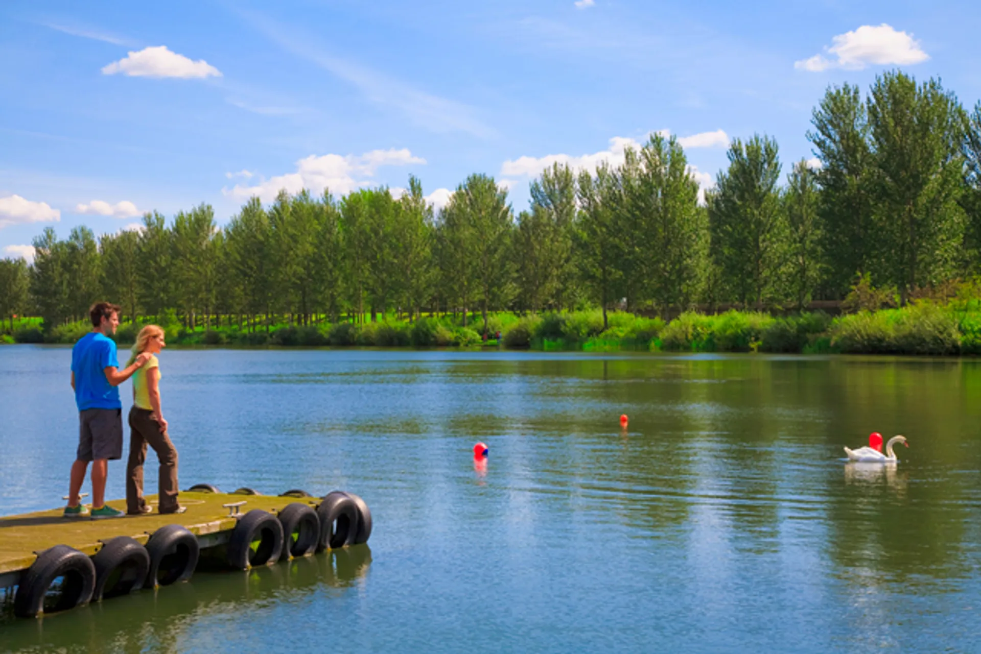 The image shows two people standing on a jetty by a calm body of water