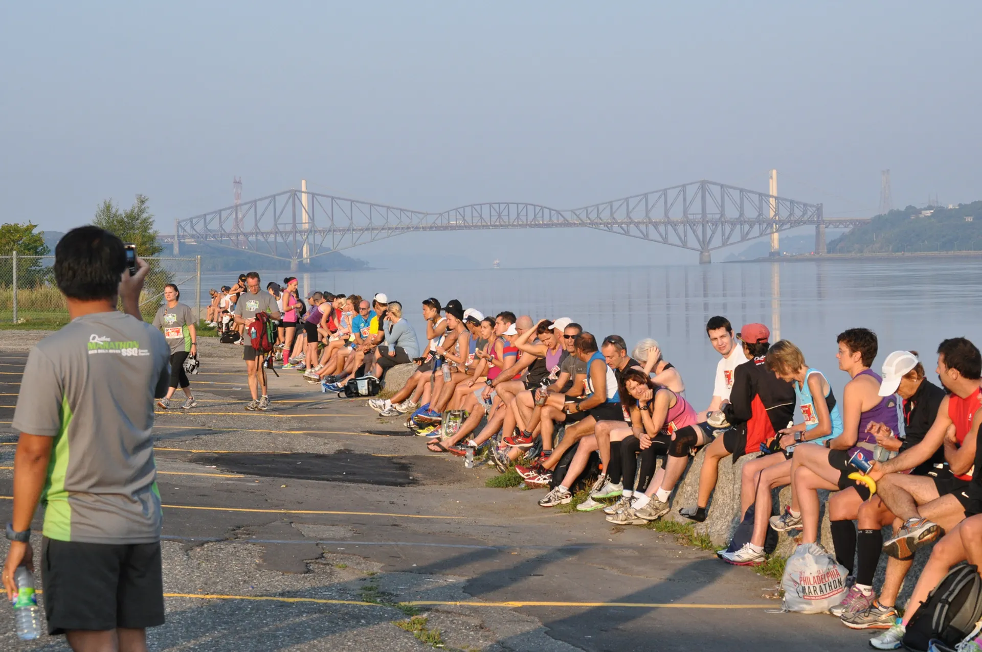 The image features a group of people gathered along a waterfront, with a large bridge