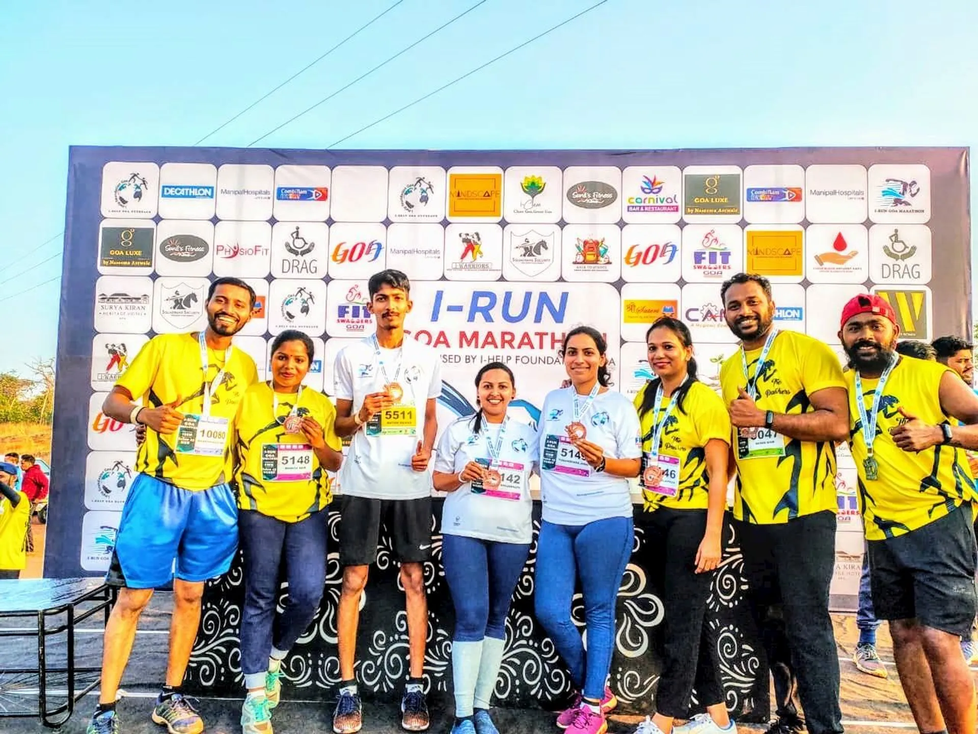 The image shows a group of people posing in front of a backdrop with various logos. It seems to be a marathon or running event, as indicated by their attire and race bibs. They're all smiling and appear to be celebrating their participation or achievement.