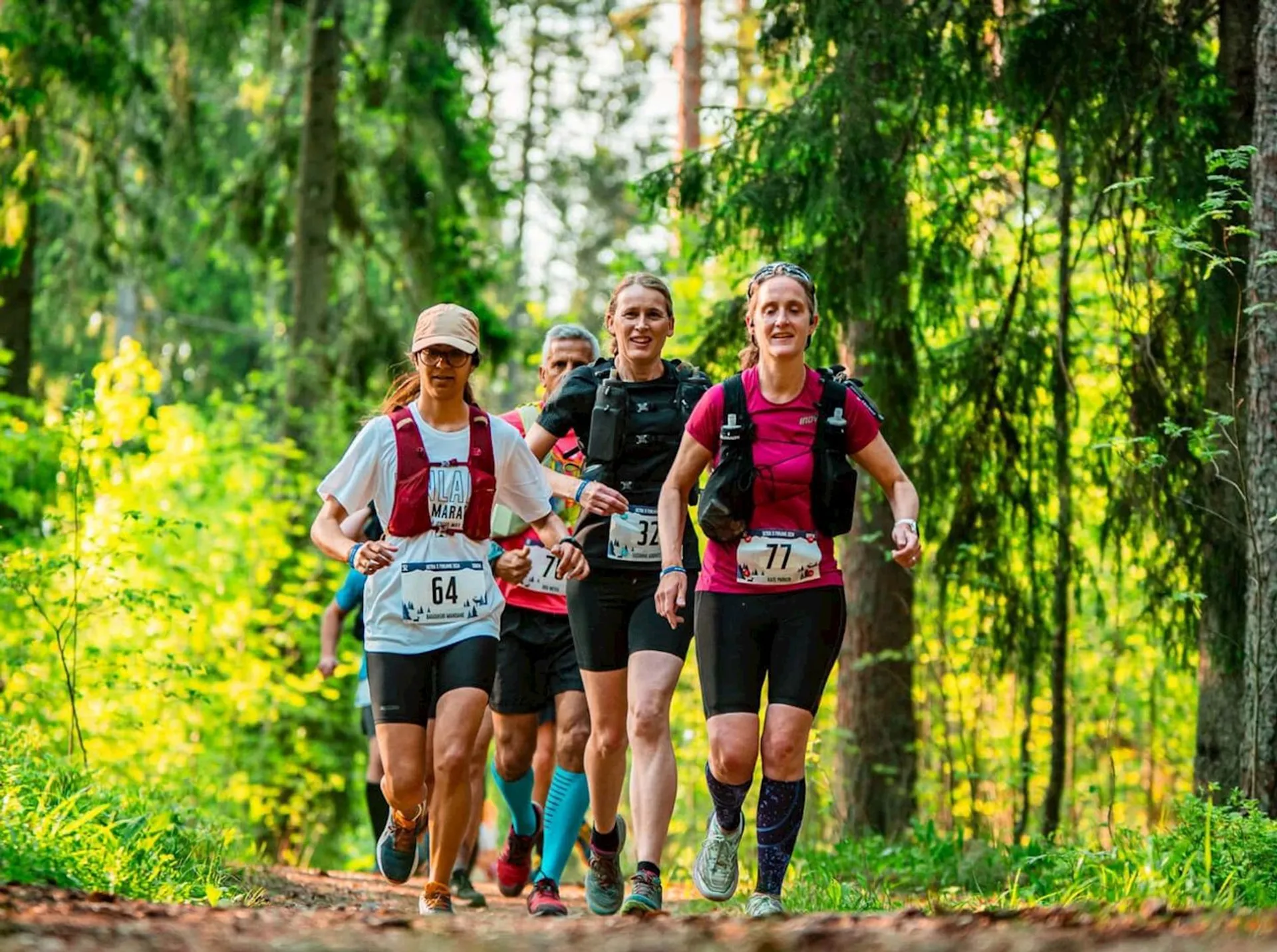 The image shows a group of people running together along a forest trail. They appear to be participating in a trail running event, as they are wearing athletic gear and bib numbers. The surrounding area is lush with greenery, indicating it's likely spring or summer. The runners seem to be enjoying the activity in a natural, wooded setting.