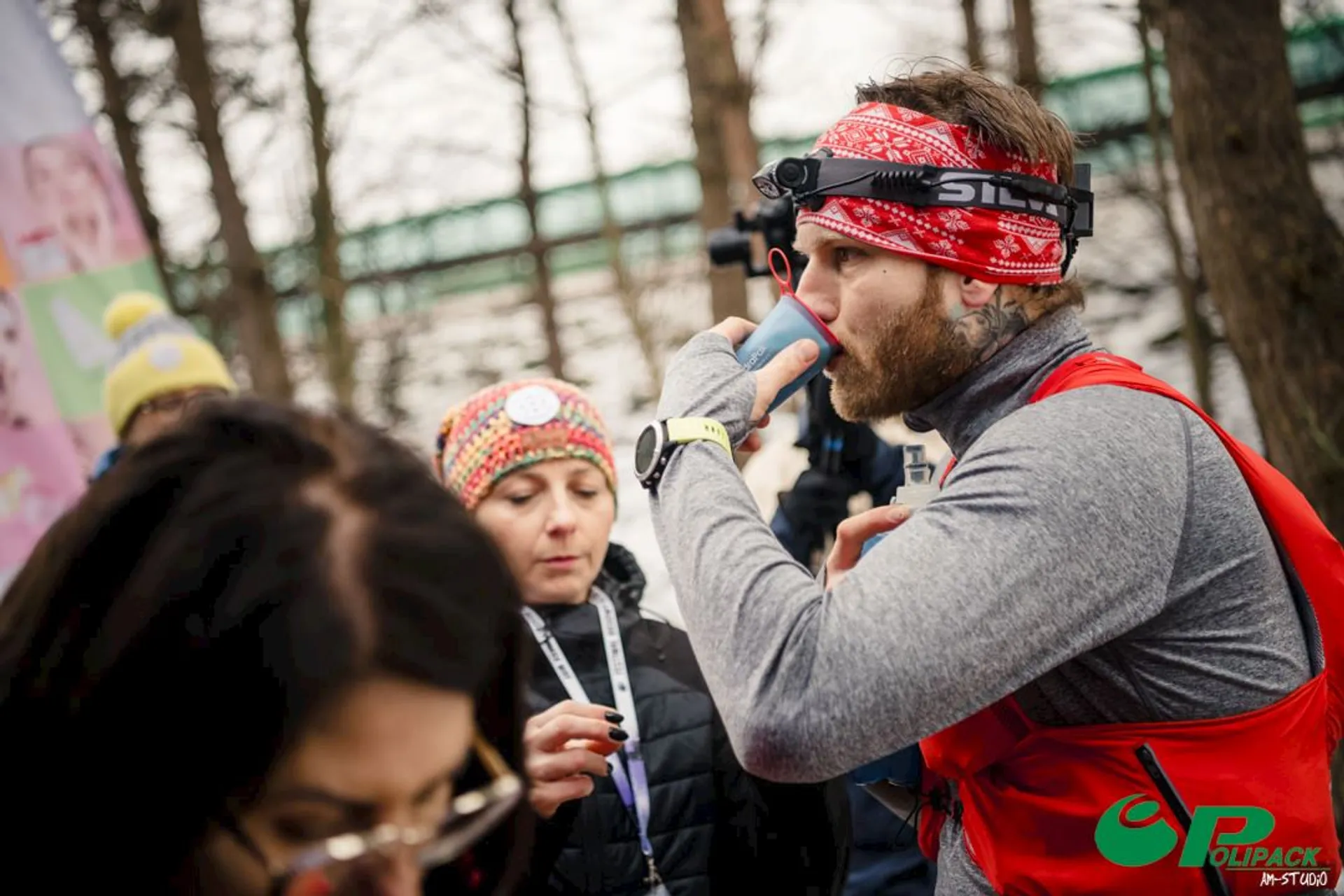 The image shows a man wearing athletic gear, including a red vest and headband, using an inhaler. There are other people around him, likely in an outdoor setting, possibly during an event or race.