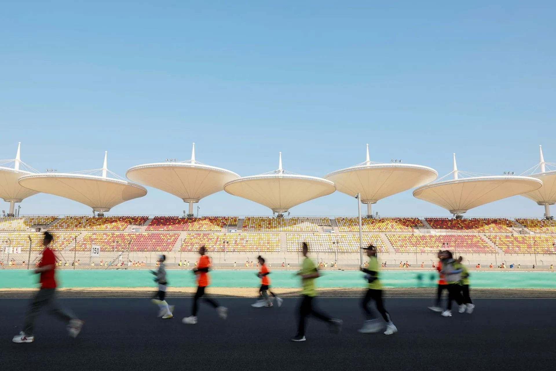 The image shows people running on a track in front of large, uniquely designed stadium structures with umbrella-like roofs. The setting suggests it could be a sports event or marathon, with the stadium and seating in the background.