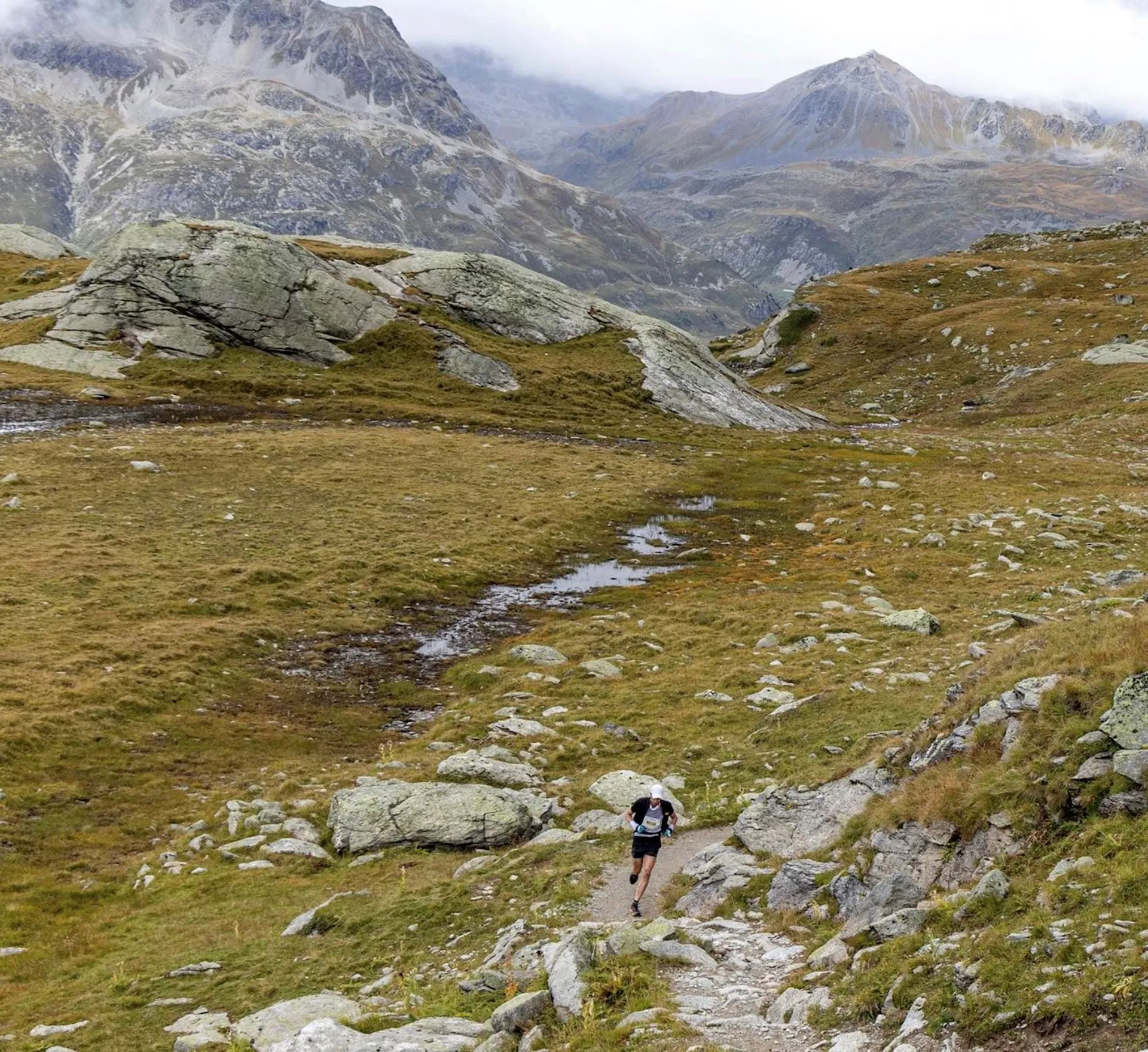 The image shows a person hiking on a trail in a mountainous area. The person is wearing a backpack and appears to be moving along a dirt path. The surrounding landscape is comprised of rolling hills with grass and scattered rocks. The mountains in the background rise steeply and seem to have some patches of snow or rock on their surfaces. There are some clouds hanging low, suggesting that the weather might be cool or that the altitude is high enough to be in the clouds. This appears to be a serene, natural environment ideal for hiking and outdoor activities.