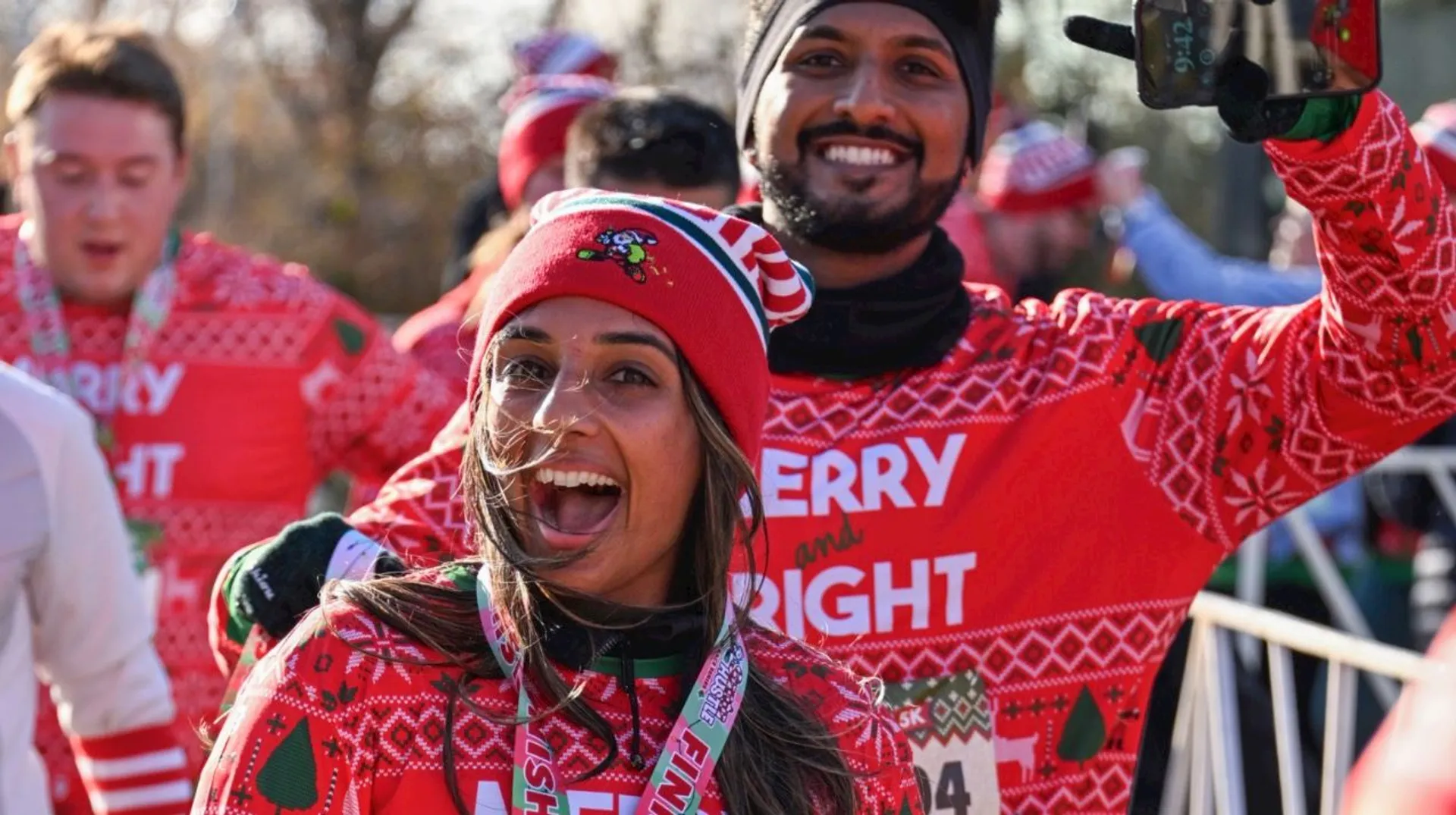 The image shows people participating in a festive running event. They are wearing matching holiday-themed attire with phrases like "Merry and Bright" and have medals around their necks, indicating they have completed a race. The atmosphere appears joyful and celebratory.