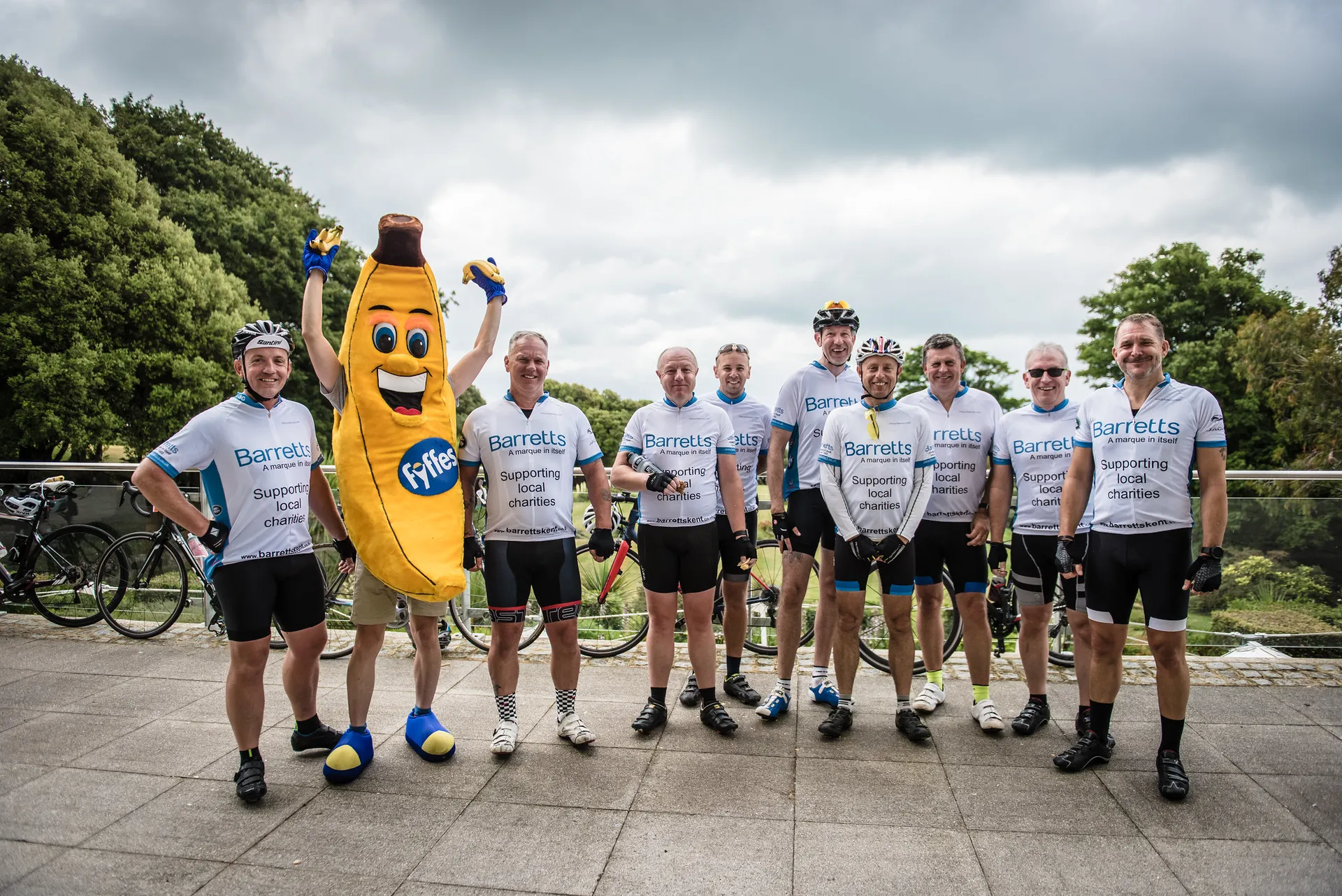 The image features a group of individuals dressed in cycling gear, posing for a photo together outdoors. They all seem to be wearing matching jerseys with the word "Barretts" on them, suggesting they might be part of a team or participating in a cycling event sponsored by an entity with that name. Additionally, there's a person in the center dressed in a mascot costume resembling a banana, bringing a whimsical and fun element to the photo. The setting appears to be a park or a recreational area with trees and a cloudy sky in the background. Some of the individuals are wearing bicycle helmets and sunglasses, sporting the look of recreational or amateur cyclists.