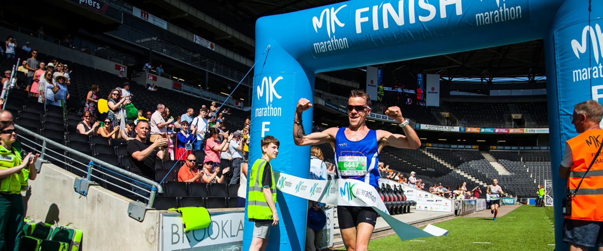 The image shows a runner crossing the finish line at a marathon event. There is a crowd of spectators in the background cheering, and event staff are present. The finish line is marked with an archway that has "MK marathon" and "FINISH" written on it. The runner appears to be celebrating the completion of the race.