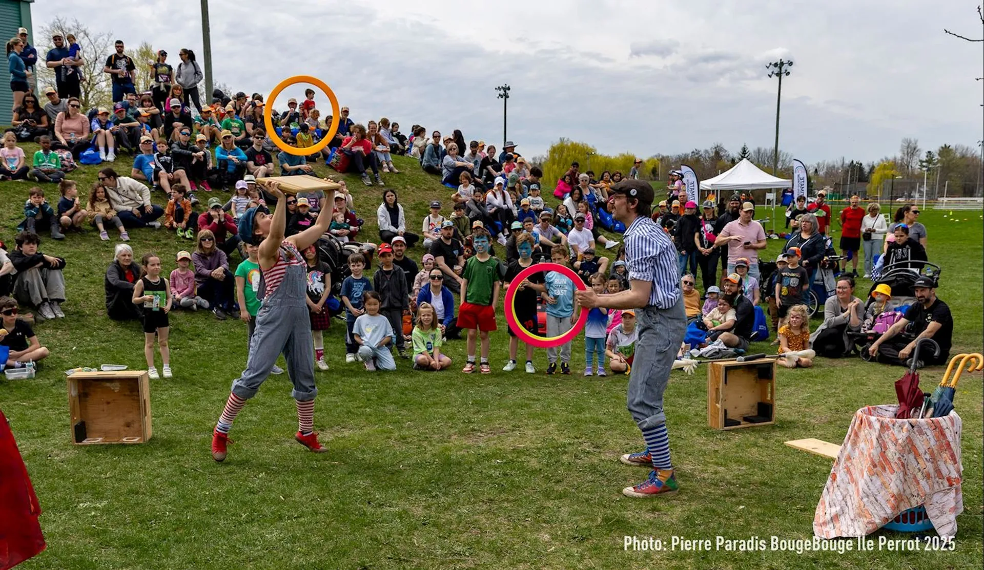 The image shows a festive outdoor event with two performers engaging in a circus act in front of a seated and standing audience on a grassy area. The performers are juggling rings and wearing playful costumes. Children and adults are watching the performance, and there's a lively, picnic-like atmosphere.