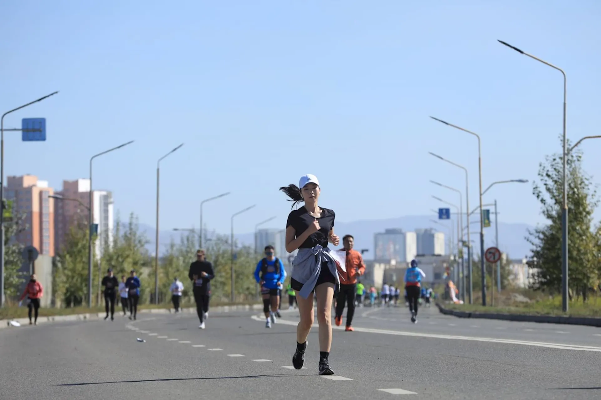The image shows a group of people running on a wide, empty street. There are streetlights along the sides of the road, and buildings can be seen in the background. This appears to be an outdoor race or marathon under a clear blue sky.