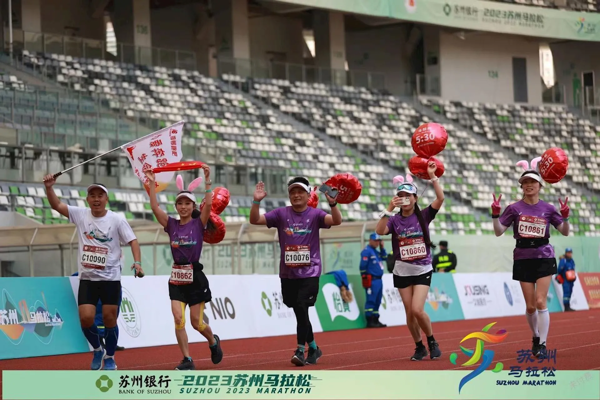 The image shows a group of runners at a marathon event. They are running on a track inside a stadium, and some are holding red balloons and a flag. The banner at the bottom indicates this is the Suzhou 2023 Marathon. The participants appear to be celebrating or finishing the race.