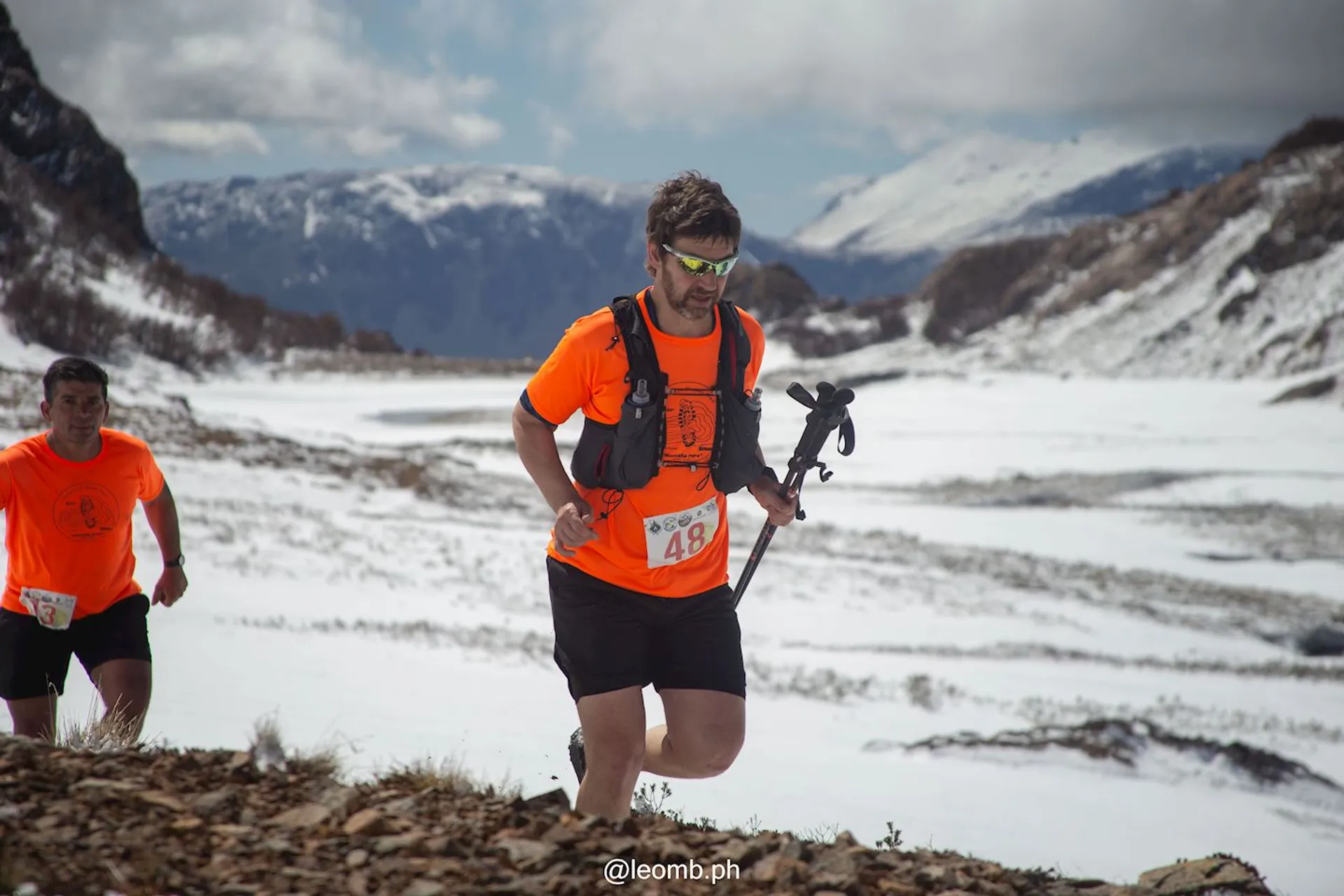 The image shows two people running in a snowy mountainous landscape. They are wearing bright orange shirts and race bibs, indicating they might be participating in a trail running event. One person is holding a pair of trekking poles. The background features snow-covered peaks and a partly cloudy sky.