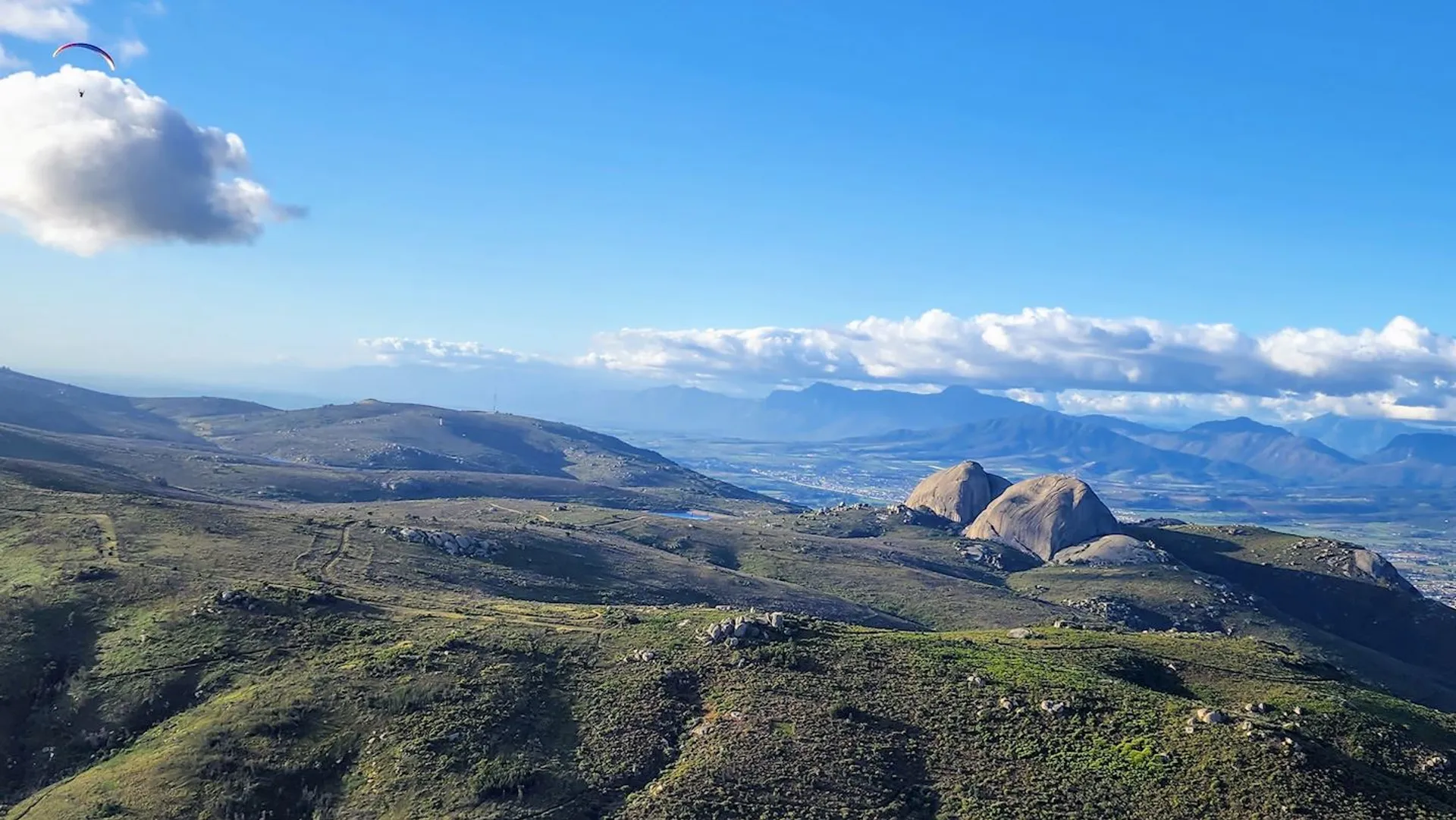 The image depicts a sprawling landscape view. In the foreground, there are rolling hills covered with greenery, possibly grasses or low-lying shrubs. Notably, there is a large boulder or rock formation prominently featured in the mid-ground, which appears quite distinctive due to its size and isolated position on the hillside. The background reveals more mountain ranges layered in the distance, suggesting a vast, open natural environment.

The sky above is mostly clear with a few clouds, and the lighting conditions indicate either early morning or late afternoon, with sunlight casting shadows and giving the landscape a warm tone. This could be a scene from a rural area, possibly a national park or nature reserve, given the lack of urban development in the vista. The overall