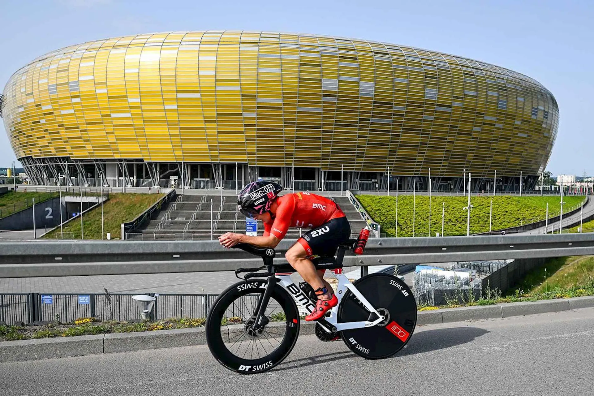 The image shows a cyclist in racing gear riding a time trial bicycle on a road. In the background, there is a large, oval-shaped building with a distinctive yellow and gold exterior. The cyclist appears focused and aerodynamic, typical of a time-trial position in cycling.