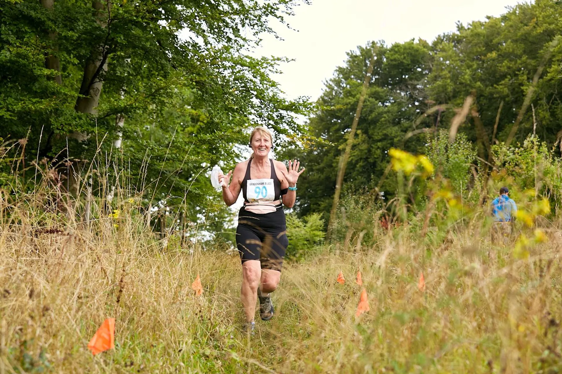 The image shows a person running along a trail through a grassy area. They are participating in a race, as indicated by the bib number on their shirt. The person appears to be smiling and waving at the camera. There are trees in the background and some orange markers on the ground beside the trail.