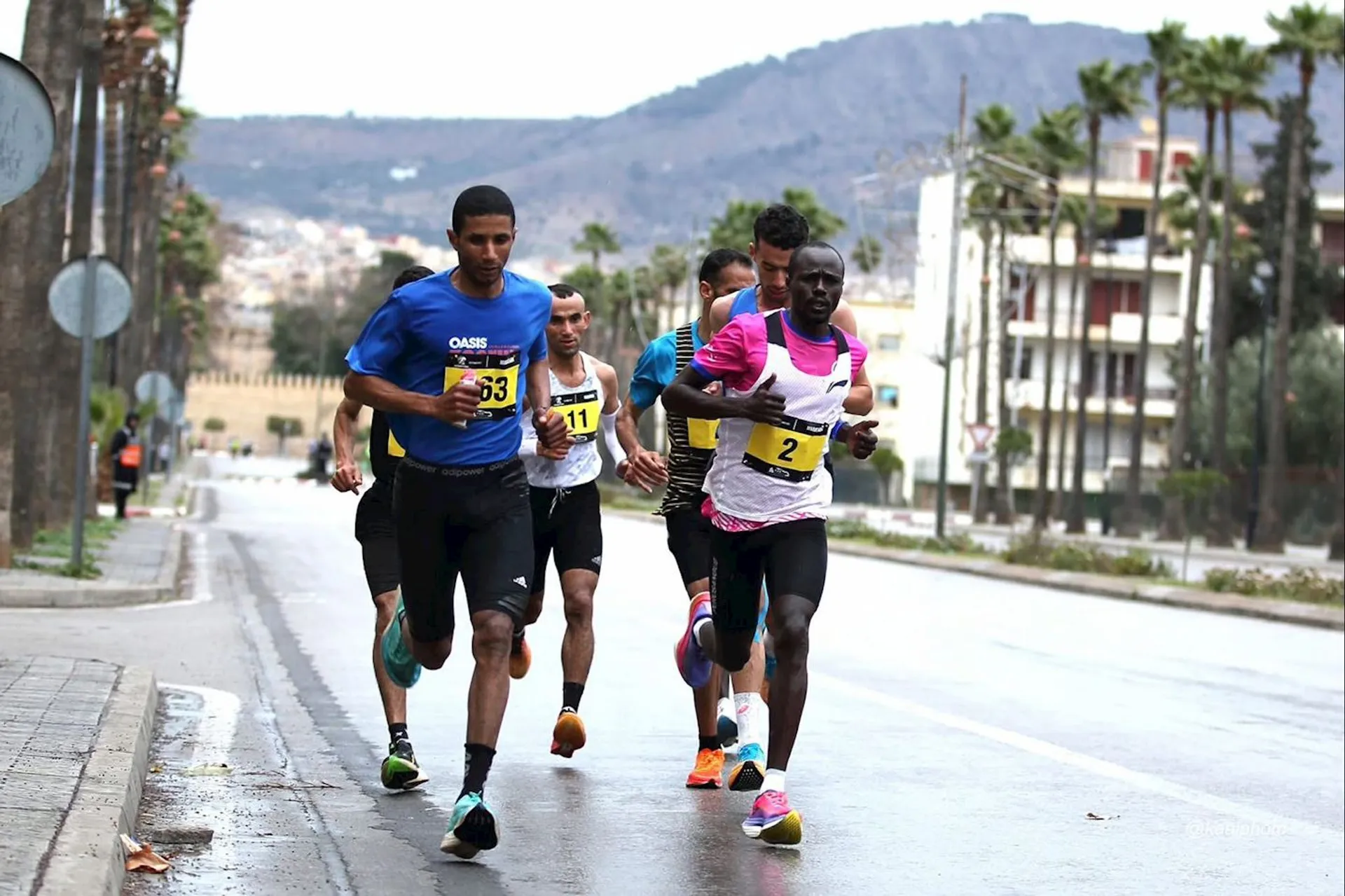 The image shows a group of runners participating in a road race. They are running on a street with numbered race bibs on their shirts. The background features buildings, a mountain, and trees lining the road.