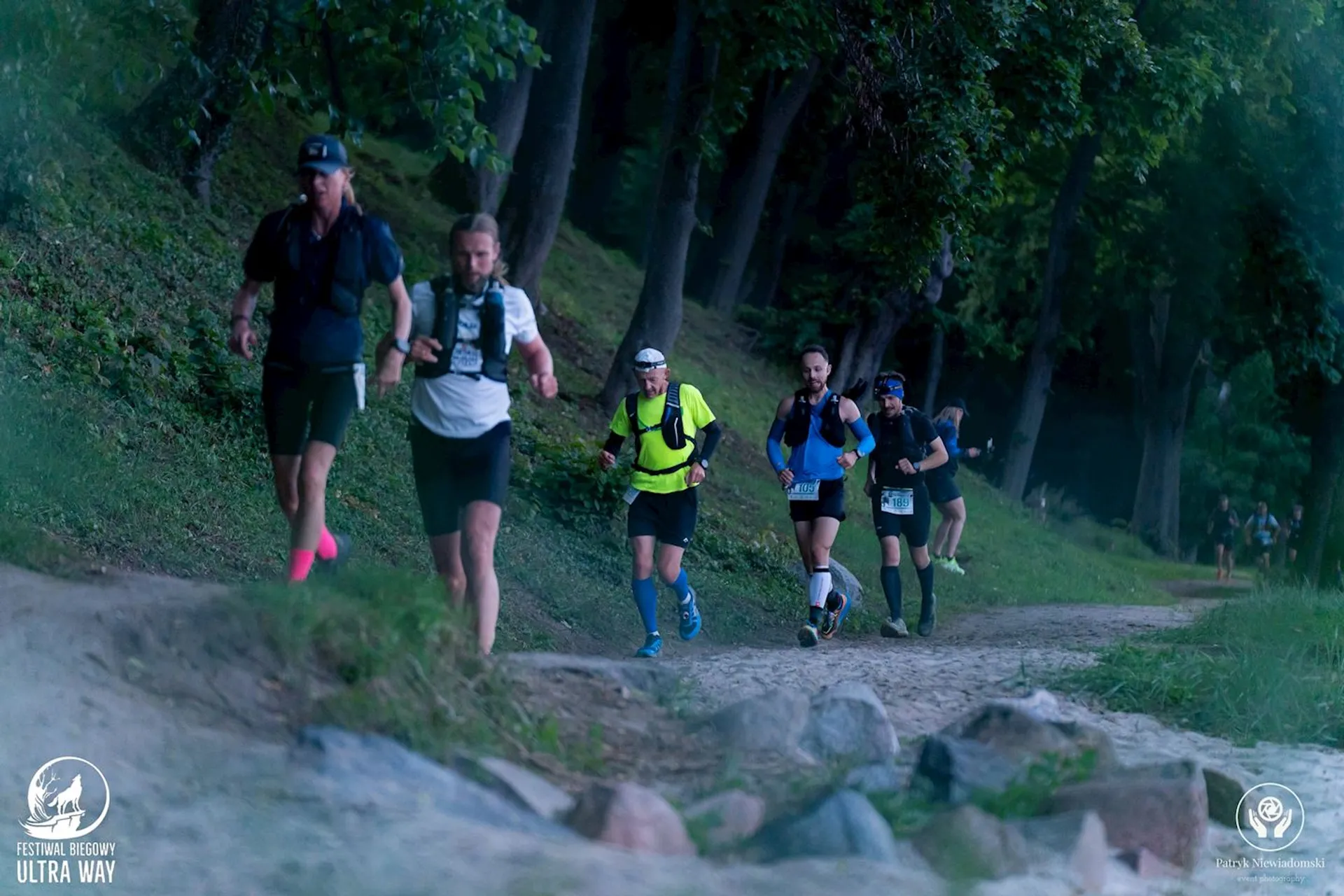 The image shows a group of people running on a trail through a wooded area. They appear to be participating in a trail or ultra-marathon event. Some are wearing running gear with hydration packs. There are logos on the image, suggesting it's part of an organized race or festival.