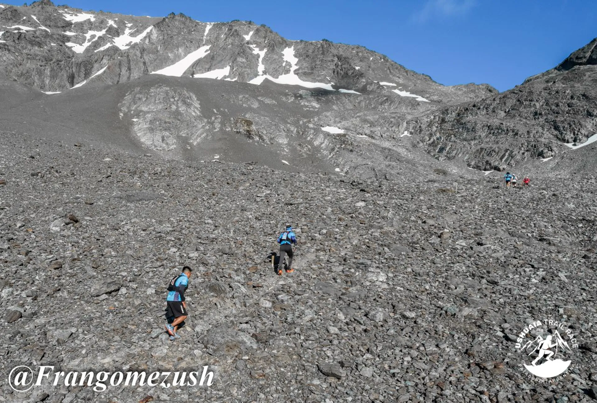 The image shows several people hiking up a rocky, mountainous terrain. There are patches of snow on the higher slopes, and the area appears to be rugged and challenging. The hikers are wearing outdoor gear suitable for a mountainous environment. The scene is set under a clear blue sky.