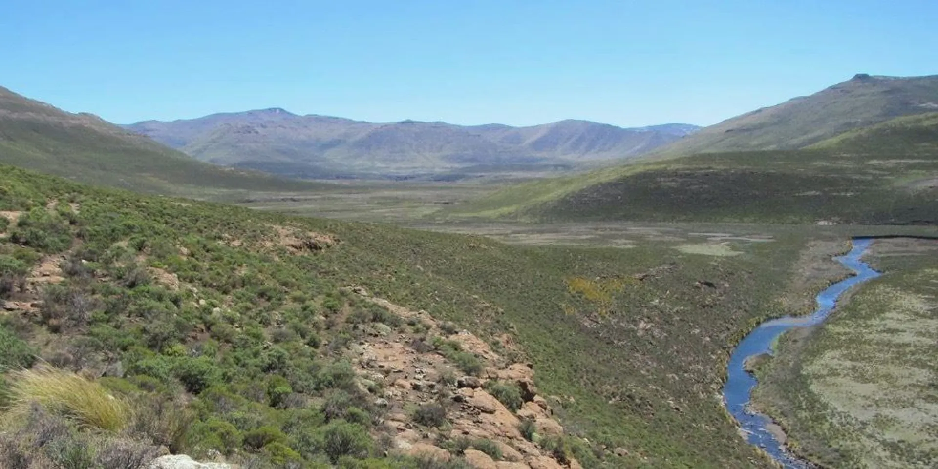 The image shows a wide, open landscape with rolling hills and a river meandering through the valley. The land is covered with grasses and shrubs, and there are mountains in the background. The sky is clear and blue, suggesting a bright, sunny day.