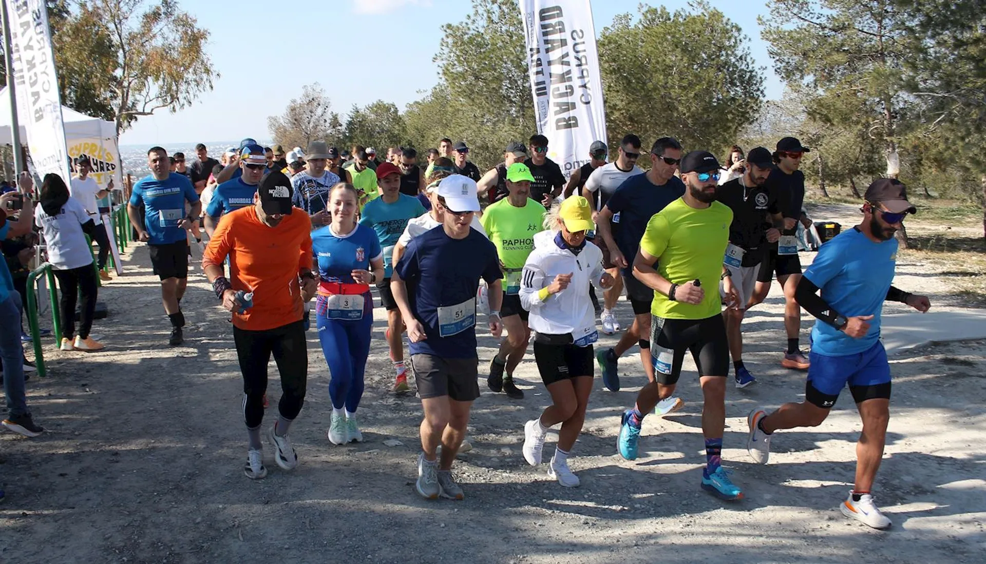 The image shows a group of people running in an outdoor race or marathon. The participants are wearing athletic clothing and shoes, and each runner has a bib number pinned to their shirts. Some spectators can be seen in the background. The setting appears to be a sunny day with trees lining the path.