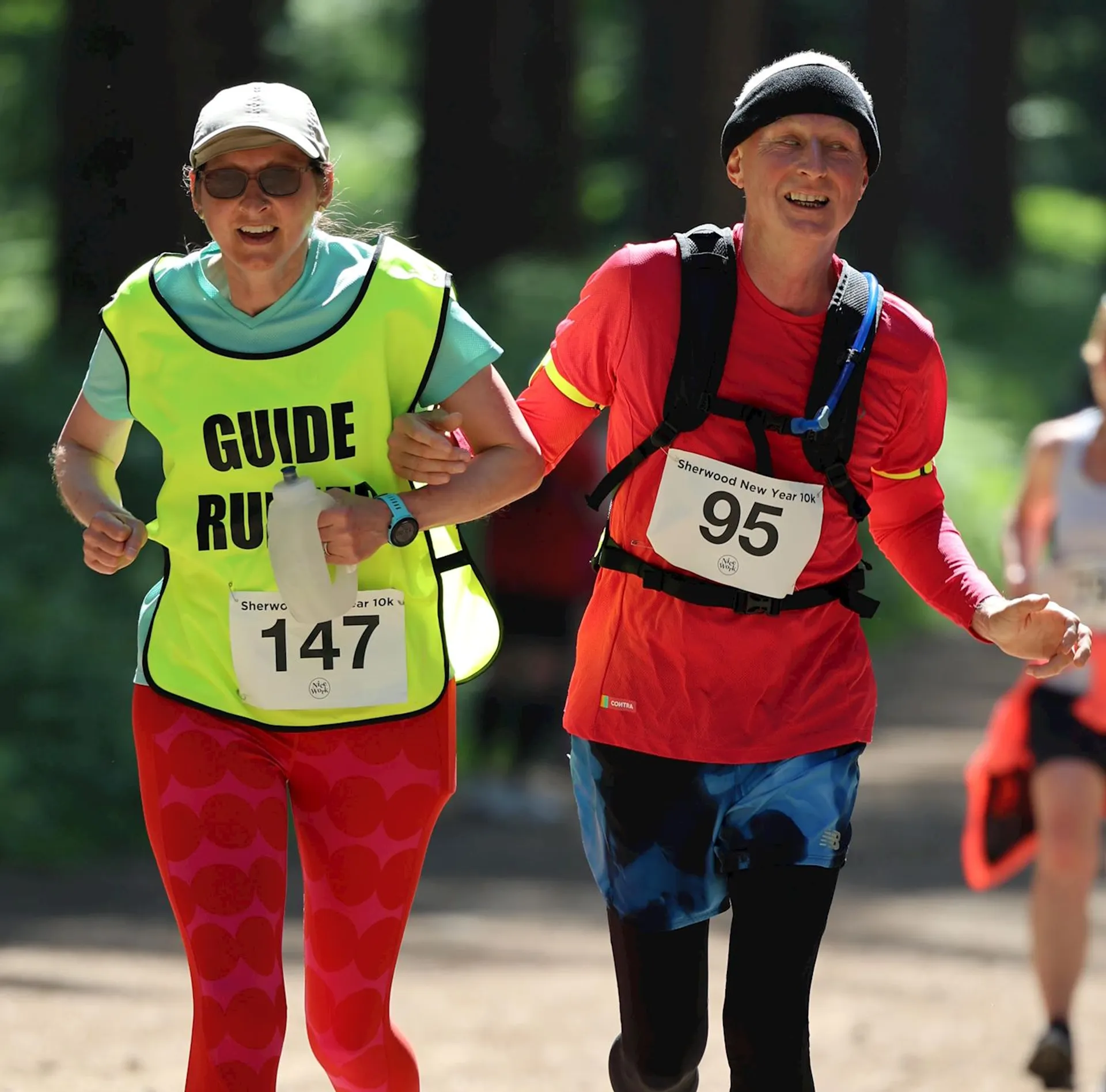 The image shows two people engaged in a running event. The person on the left is wearing a bright yellow bib marked "GUIDE RUNNER" along with a race number, suggesting they are acting as a guide for the other runner. This often occurs in events where a runner is visually impaired and requires assistance to navigate the course. The other runner, on the right, is wearing a red shirt, black shorts, and a red running cap, and they also have a race number visible. Both are wearing sunglasses and appear to be running on a sunlit path with trees in the background, indicating that the event is taking place outdoors, likely in a park or forested area. They seem focused and are likely working together to maintain a steady pace during
