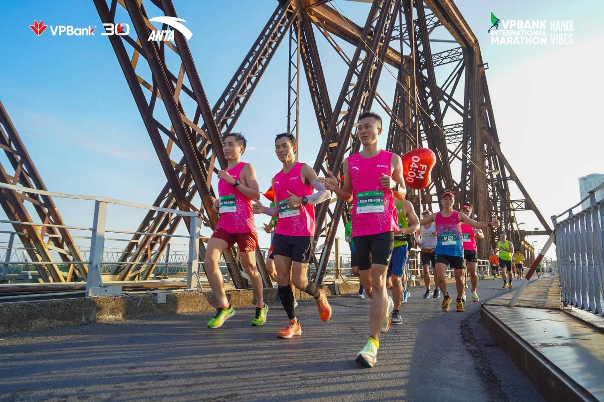 The image shows a group of runners participating in a marathon. They are running on what appears to be a bridge with a metal truss structure, which suggests it may be an iconic or historical bridge. The runners are wearing bibs, which typically display participant numbers in organized races. They seem to be in a competitive yet friendly atmosphere, indicated by their athletic attire and focused expressions.

Prominently displayed are banners with logos indicating the event is sponsored by "VPBank" and possibly related to a "Hanoi Marathon." The sky is clear, and the sun is shining, which suggests good weather for the race. The image captures the energy and determination typically seen at such sporting events.