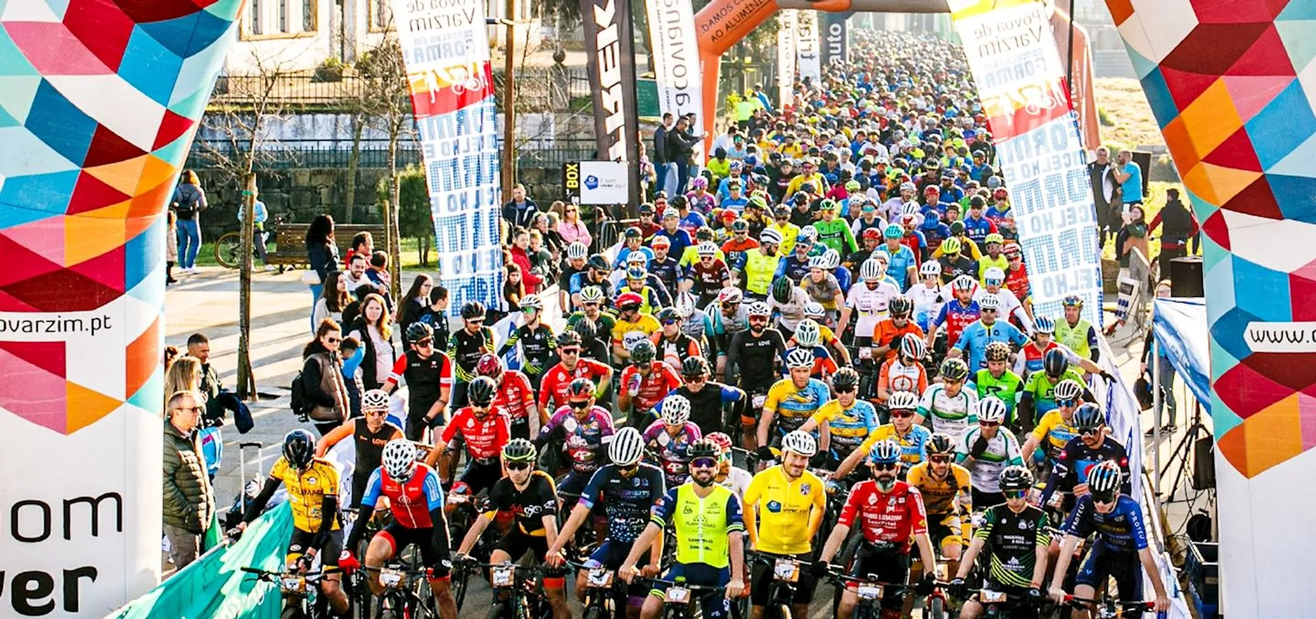 The image shows a large group of cyclists lined up at the start of a cycling event or race. They are wearing various colorful cycling outfits and helmets, indicating the presence of different teams or individual participants. There are banners with logos and advertisements, suggesting that the event might be sponsored by a variety of organizations. The background features onlookers and a cityscape, hinting that the event is taking place in an urban area. The atmosphere seems lively and competitive, typical of a sporting event kick-off.