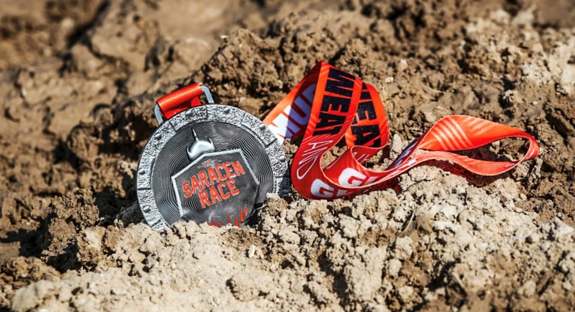 This image shows a medal resting on muddy ground. The medal is attached to a red and orange striped lanyard and has the text "SARACEN RACE" prominently displayed.