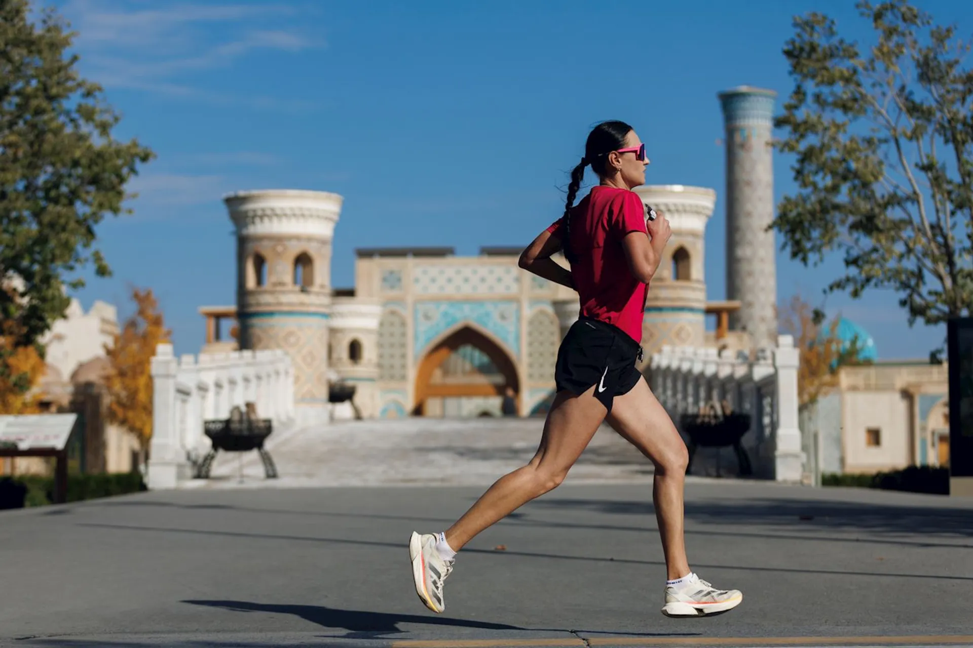 In the image, there is a person jogging in a scenic area. The runner is wearing a red shirt, black shorts, and sunglasses. In the background, there is an ornate building with blue and white tiles, featuring arches and tall cylindrical towers, which suggests a location with historical or cultural significance. The sky is clear and trees line the path.