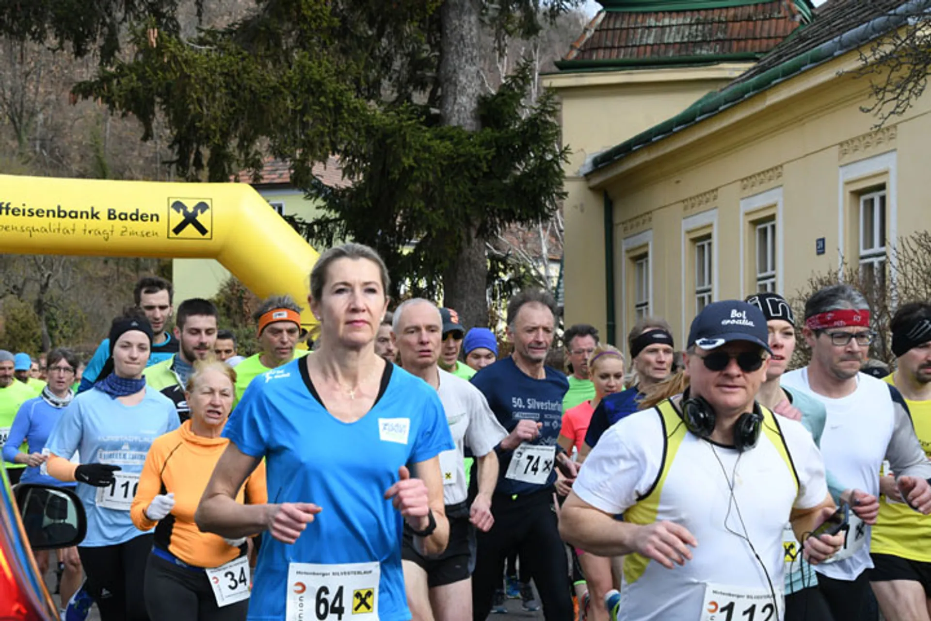 The image shows a group of people participating in a running event or race. They appear to be just at the start or early in the course, as suggested by the presence of a starting arch in the background with sponsorship branding on it. The participants are wearing running attire, including variously colored tops, running pants or shorts, and bibs with numbers on them, which is typical for organized races to help identify the runners. Some runners are wearing headphones, suggesting they might be listening to music while they run. The setting appears to be in a scenic area with trees and a building that could indicate a residential or park area. The participants have different expressions, but overall they seem focused on the race ahead.