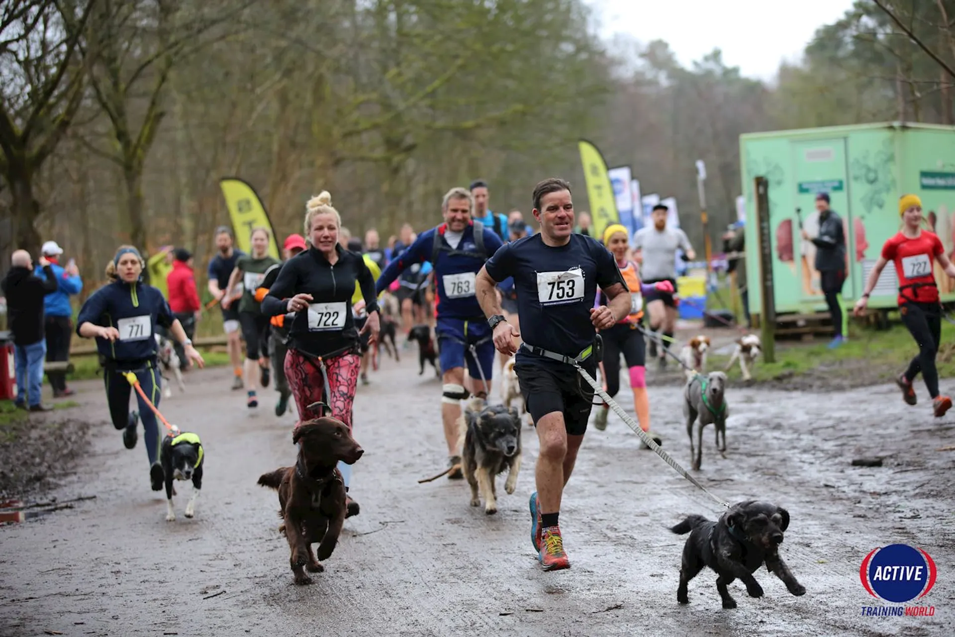 The image shows a group of people participating in a running event with their dogs. The race is taking place on a muddy path in an outdoor setting with trees and banners visible in the background. Participants have numbers on their shirts, and some are dressed in athletic gear appropriate for running. The event appears to be well-attended, with both people and dogs actively engaged in the race.