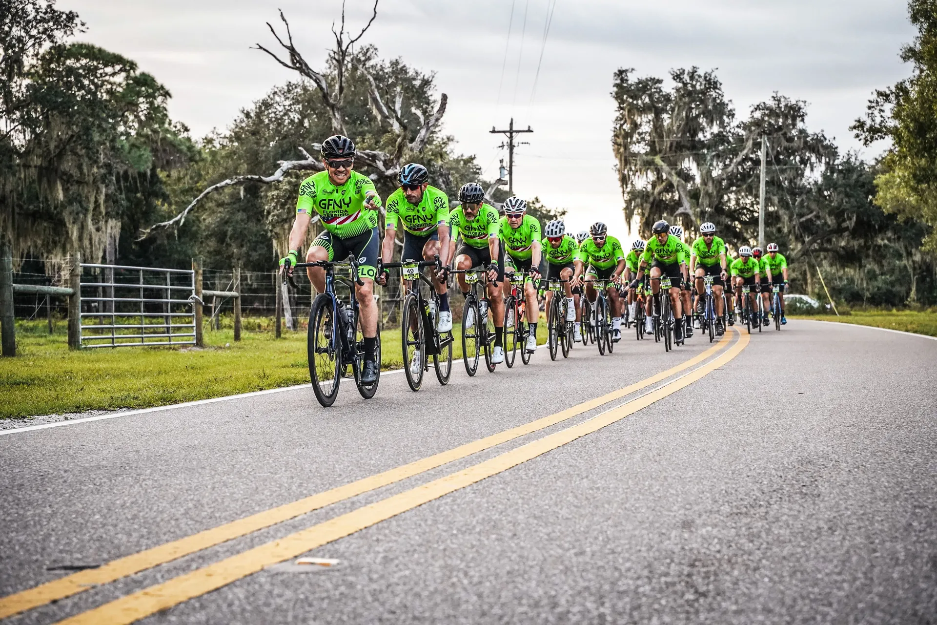 The image depicts a group of cyclists riding closely together on a road. They are
