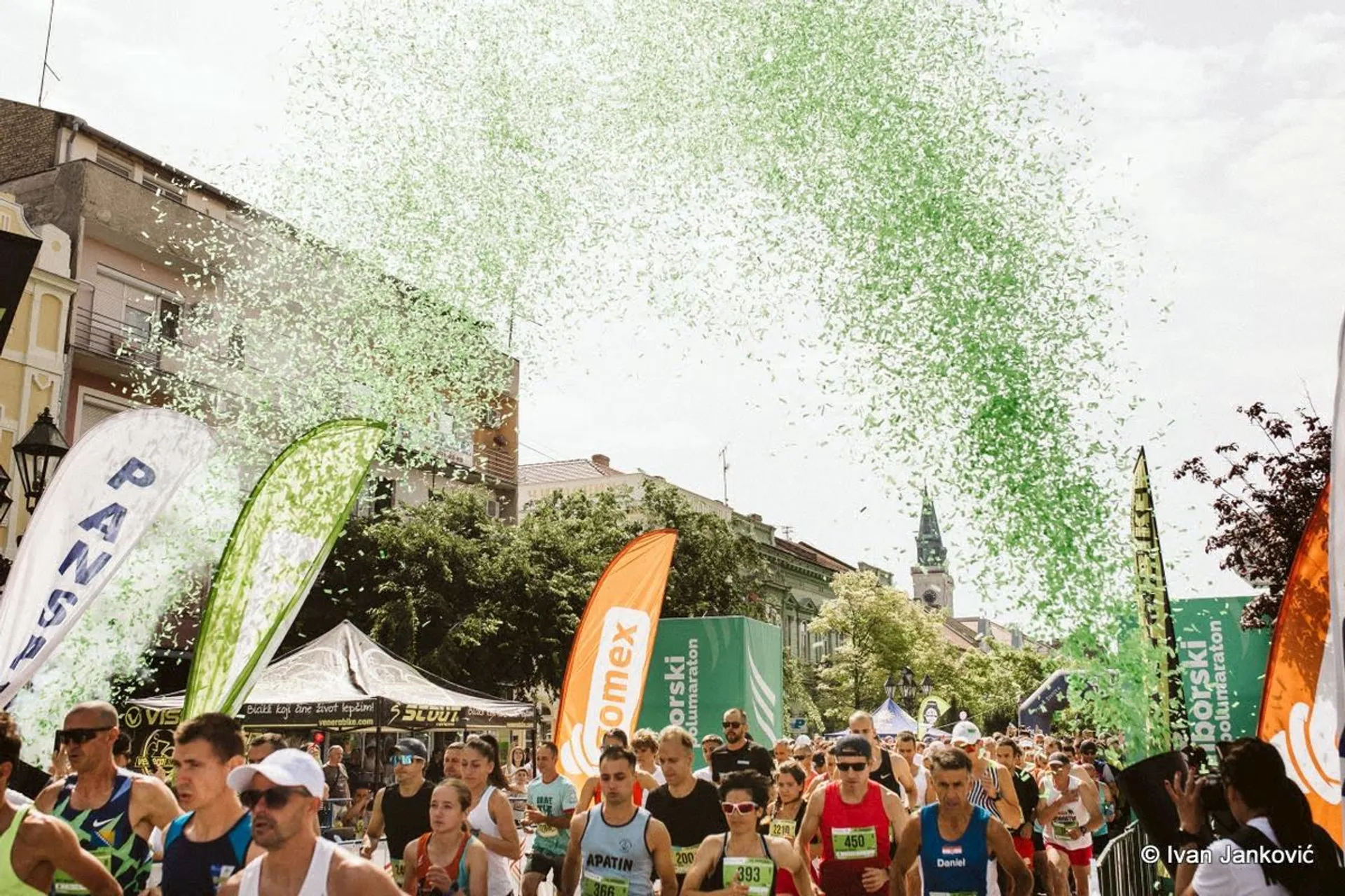The image shows a crowd of runners participating in a race. There are several banners and flags along the route, and green confetti is being launched in the air, adding to the festive atmosphere. The setting is outdoors, likely in a city with buildings visible in the background.