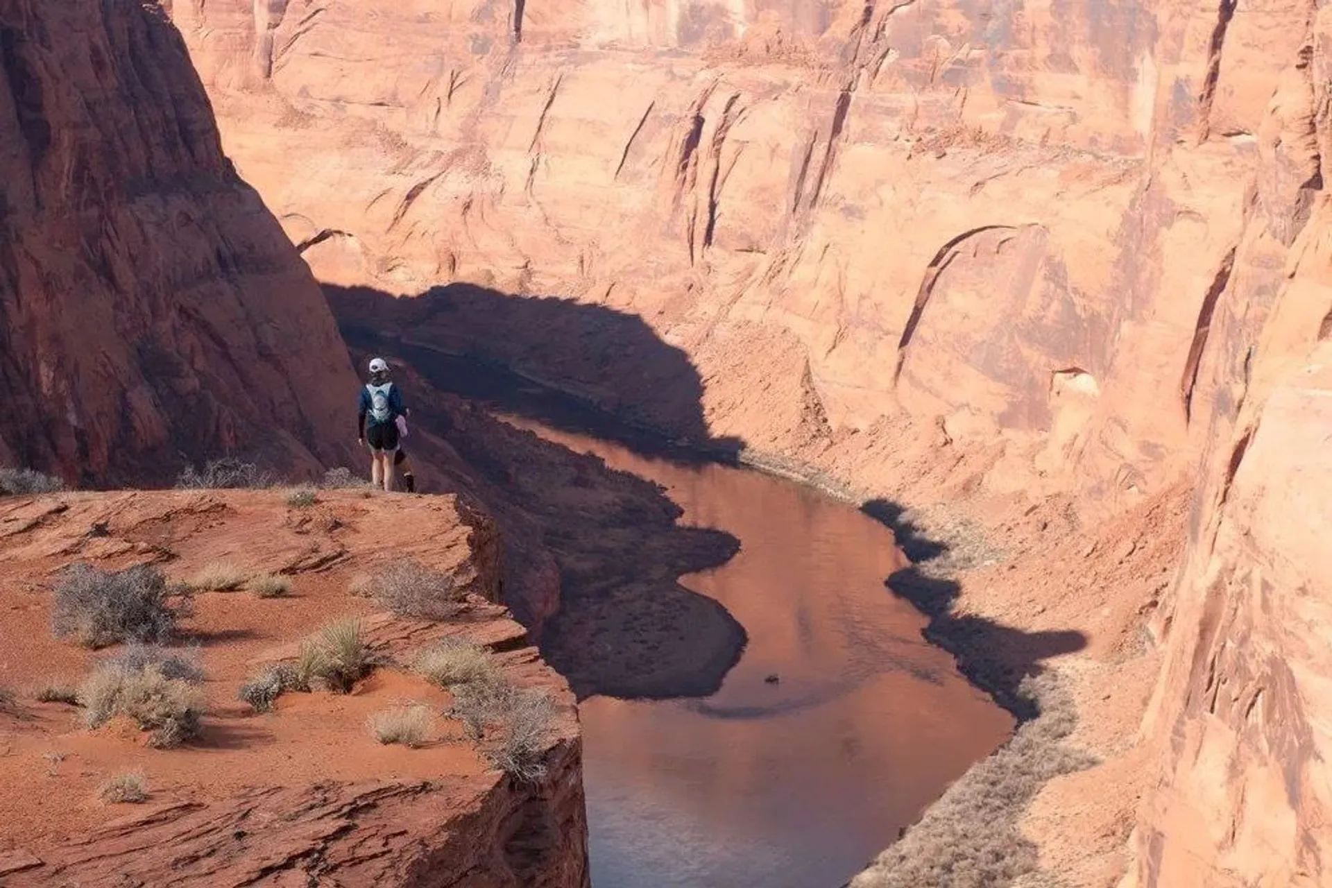 This image shows a deep canyon with steep, reddish-brown rock walls, and a river flowing through it. There is a person standing on a ledge overlooking the canyon, wearing a backpack and outdoor clothing, suggesting they are hiking or exploring the area. The landscape appears to be arid and rugged.