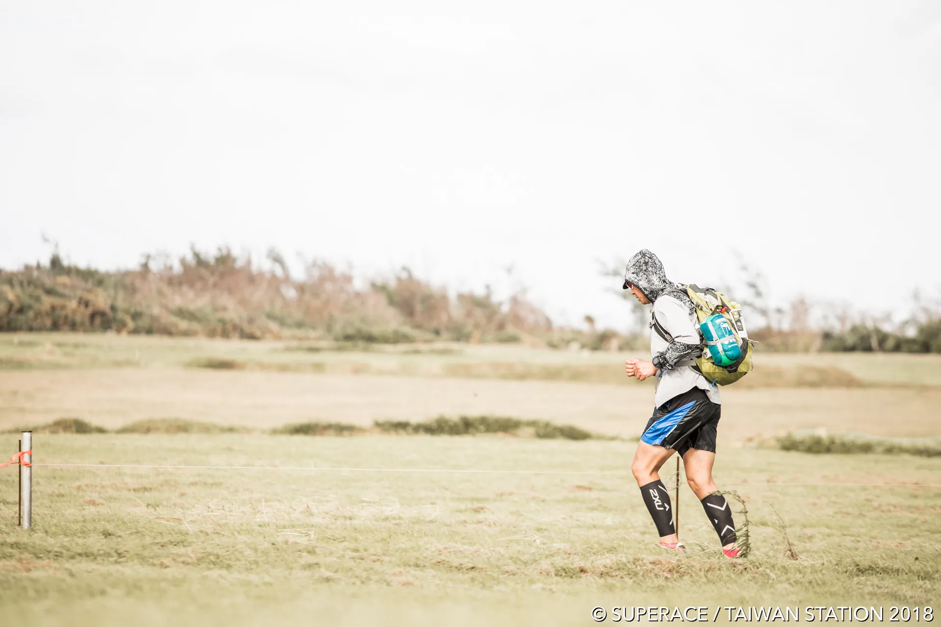 The image shows an individual who appears to be participating in an outdoor running or trekking event. The person is wearing athletic clothing suitable for such activities—shorts, leg compression sleeves, and a lightweight backpack, which is likely to be carrying hydration and other essentials. The attire also includes a hooded jacket, suggesting that weather conditions might be windy or that the runner is trying to protect themselves from the sun or other elements.

The environment looks like a wide-open field with some shrubbery and grass, indicative of a rural or natural setting that's often chosen for outdoor endurance events. The ground appears to be dry and the weather seems fair, with no immediate signs of rain or inclement conditions.

The photograph itself has a shallow depth of field,
