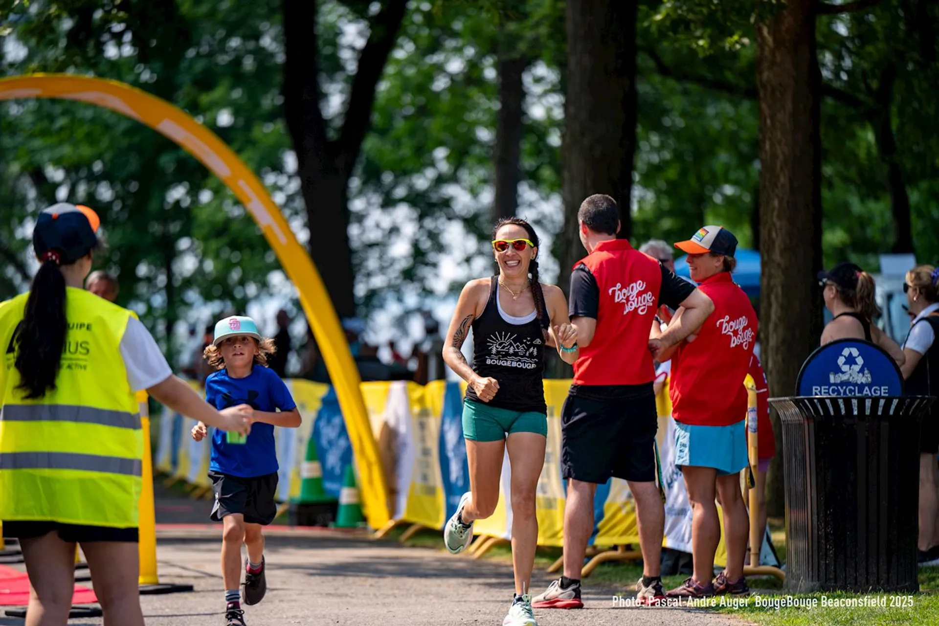 The image shows a running event taking place in an outdoor setting. A woman is running towards the finish line, smiling, while others, including volunteers and spectators, are present. There are trees and the background suggests a park or wooded area. The foreground includes a child and a person in a high-visibility vest.
