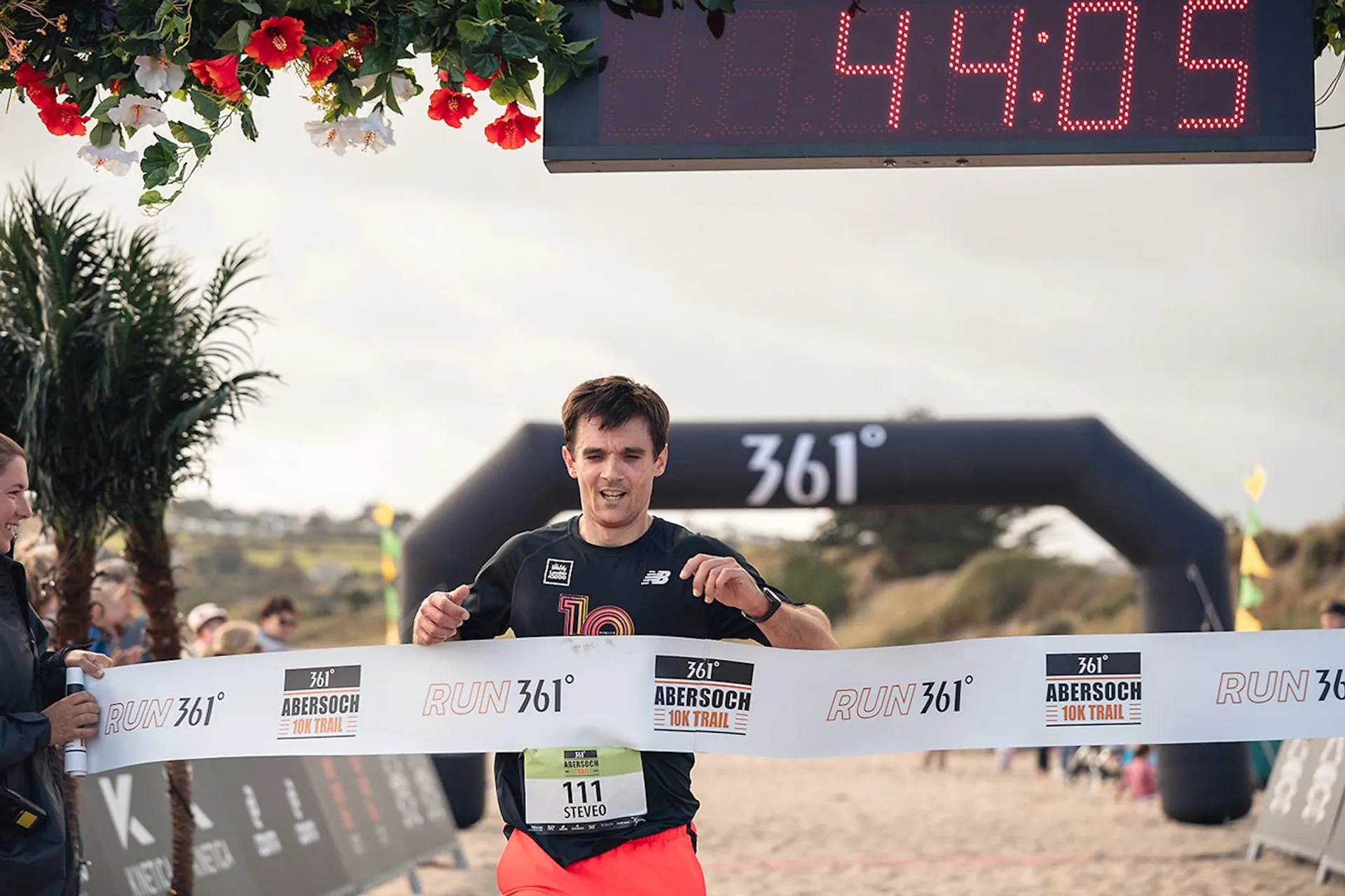 The image shows a runner crossing the finish line of a race. The setting appears to be a beach, given the sand and palm decorations. The finish line tape displays the event name "Abersoch 361° Run Trail." A digital clock above shows a time of 44:05. There are spectators on the side cheering, and signs indicating the event are visible.