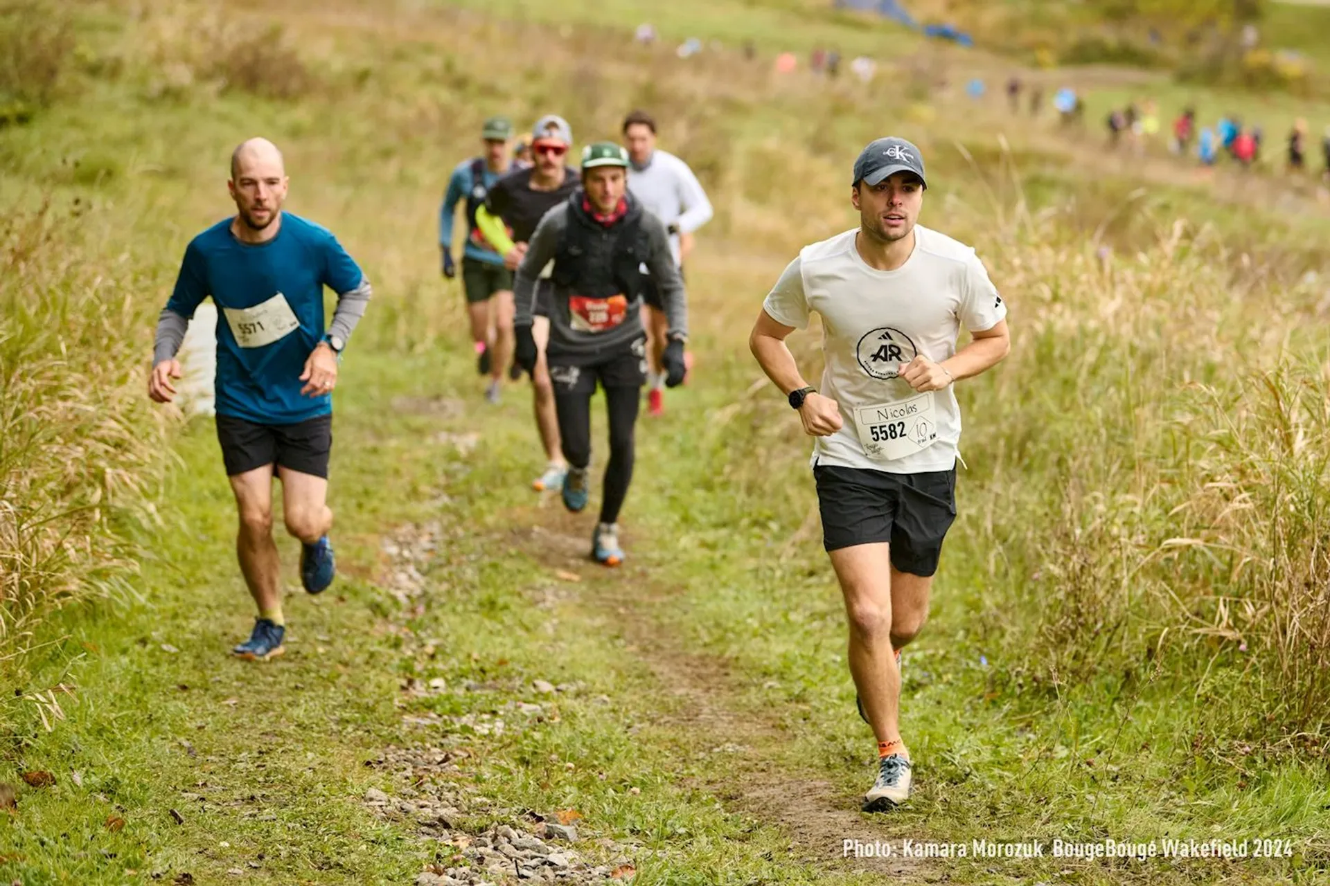 The image shows a group of people running on a trail through a grassy landscape. They appear to be participating in a trail running event. The runners are wearing athletic clothing, some with bib numbers, and the terrain looks natural and slightly rugged.