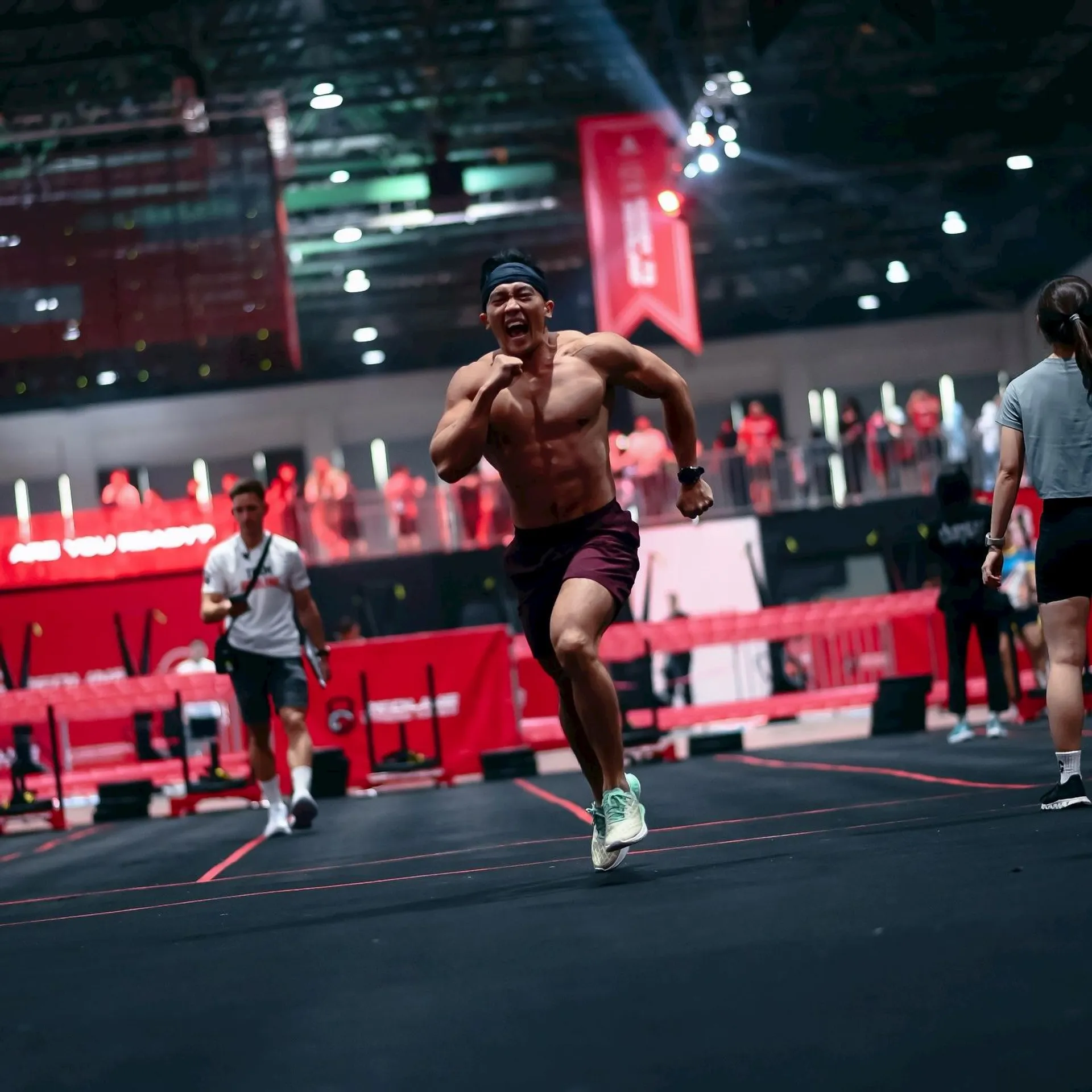 The image shows a person running indoors on a track. They are muscular and appear to be focused. There are other people and some fitness equipment in the background, along with spectators and a brightly lit area.