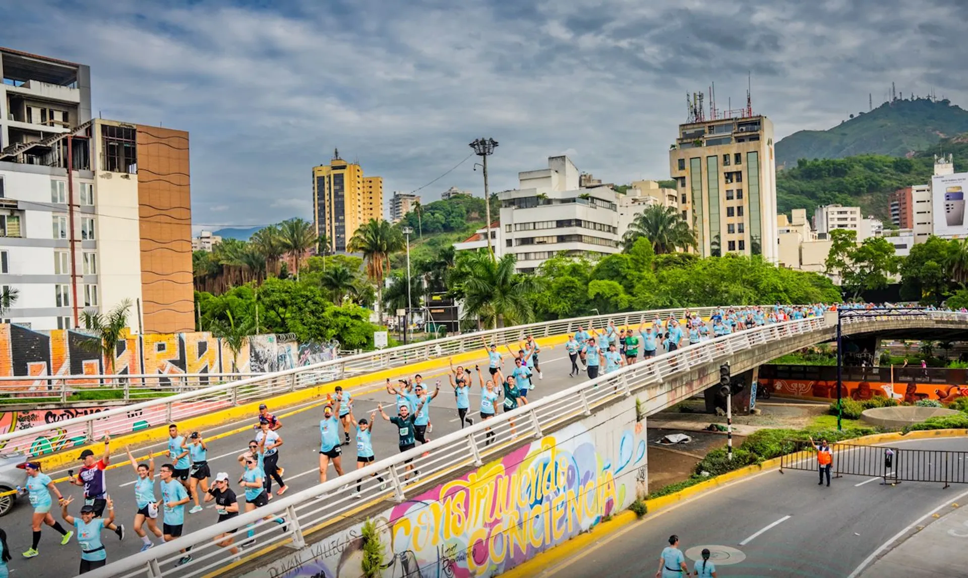 The image shows a marathon or running event taking place on an overpass in an urban area. Numerous participants are running, and some are raising their hands, likely in celebration or acknowledgment. The background features buildings, greenery, and hills or a mountain. There is graffiti on the walls along the road.