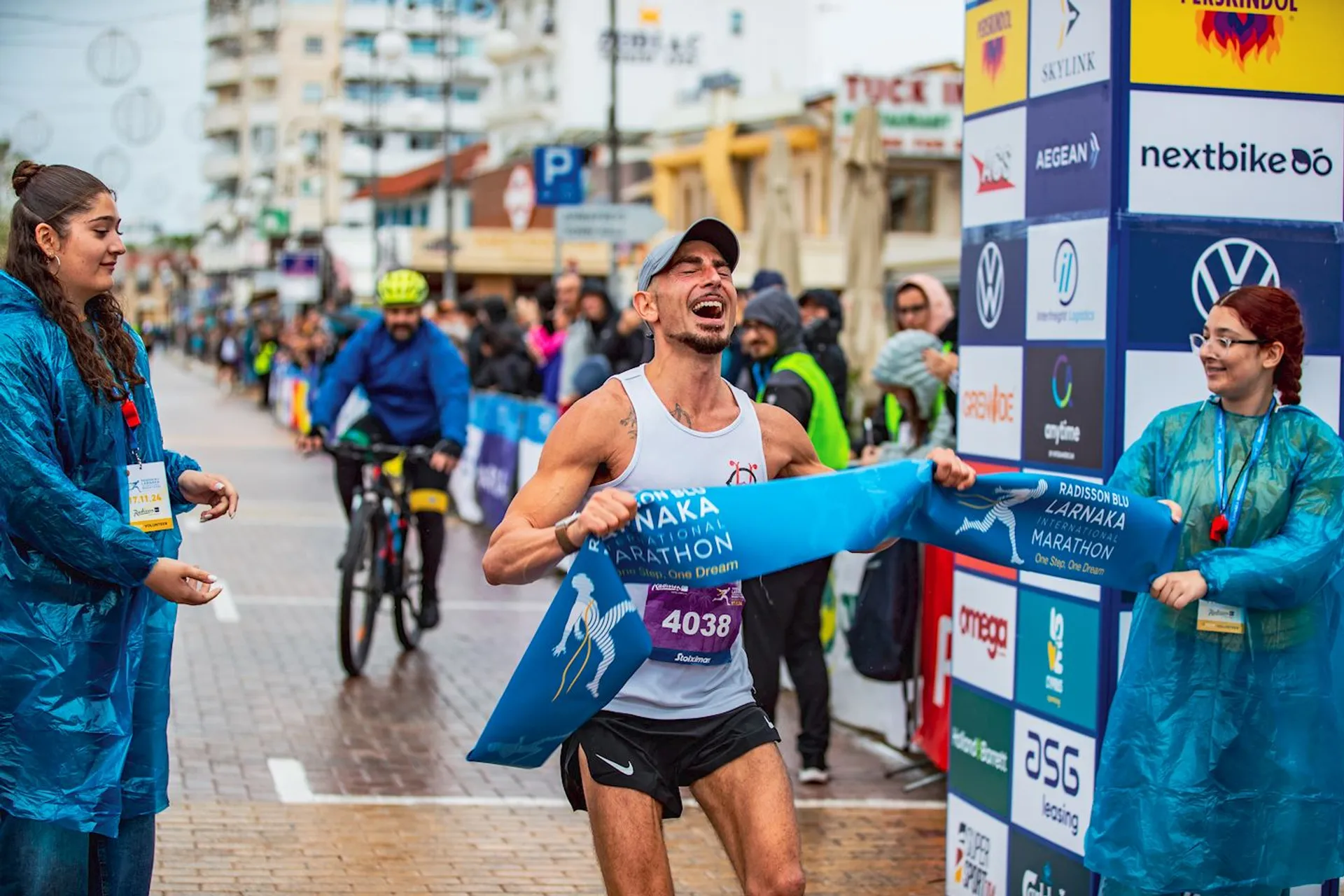 The image shows a runner crossing the finish line of a marathon. The runner looks very happy and is cheering as they break through the finish line tape. There are people around, including race officials or supporters wearing blue raincoats. The event appears to be a marathon, given the signage and setting, with sponsors' logos visible in the background.