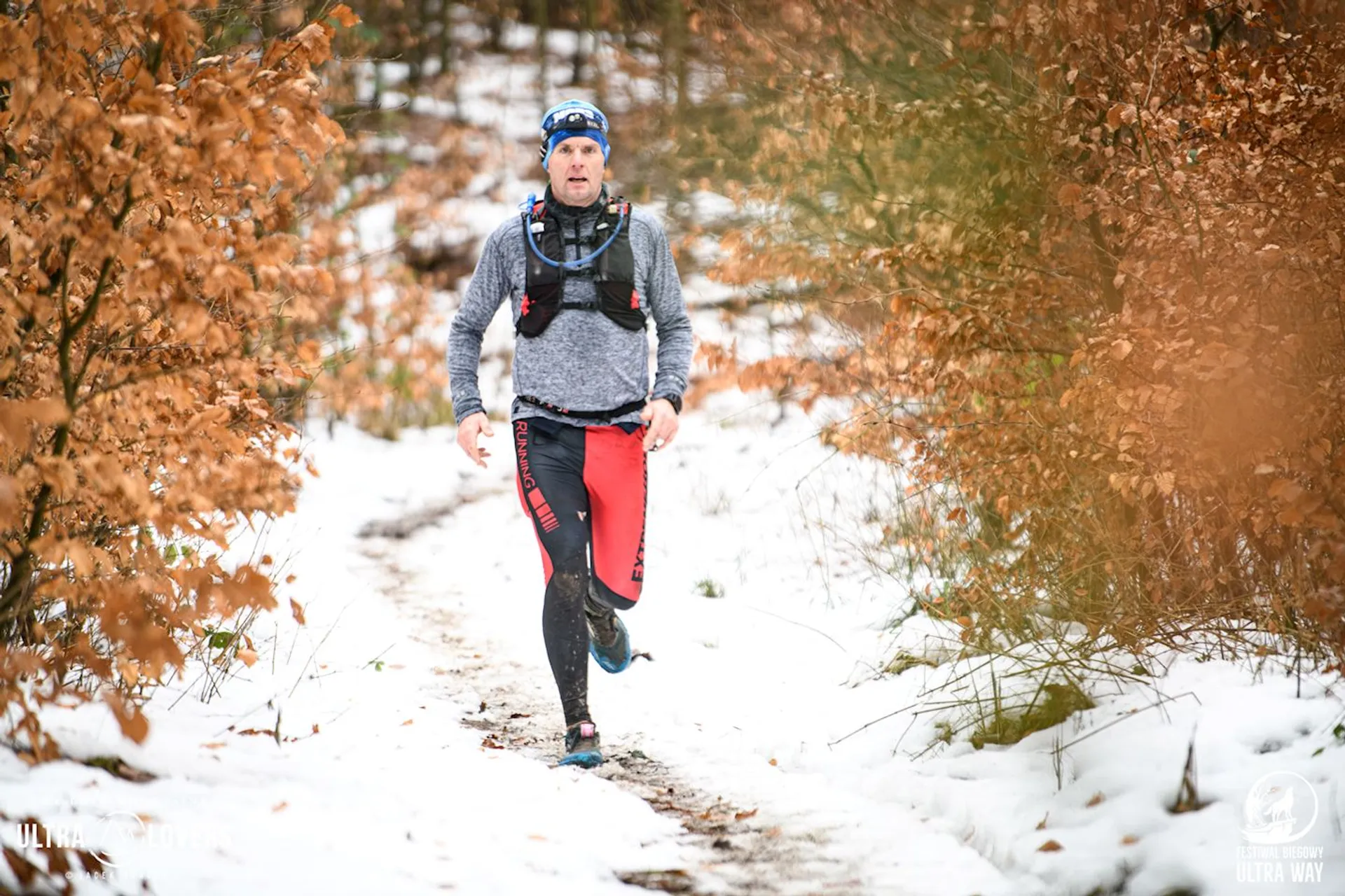 The image shows a person running on a snowy forest trail. They're wearing a gray long-sleeve top, red and black leggings, a blue headband, and a hydration vest. The surrounding trees have autumn-colored leaves, contrasting with the snow on the ground.