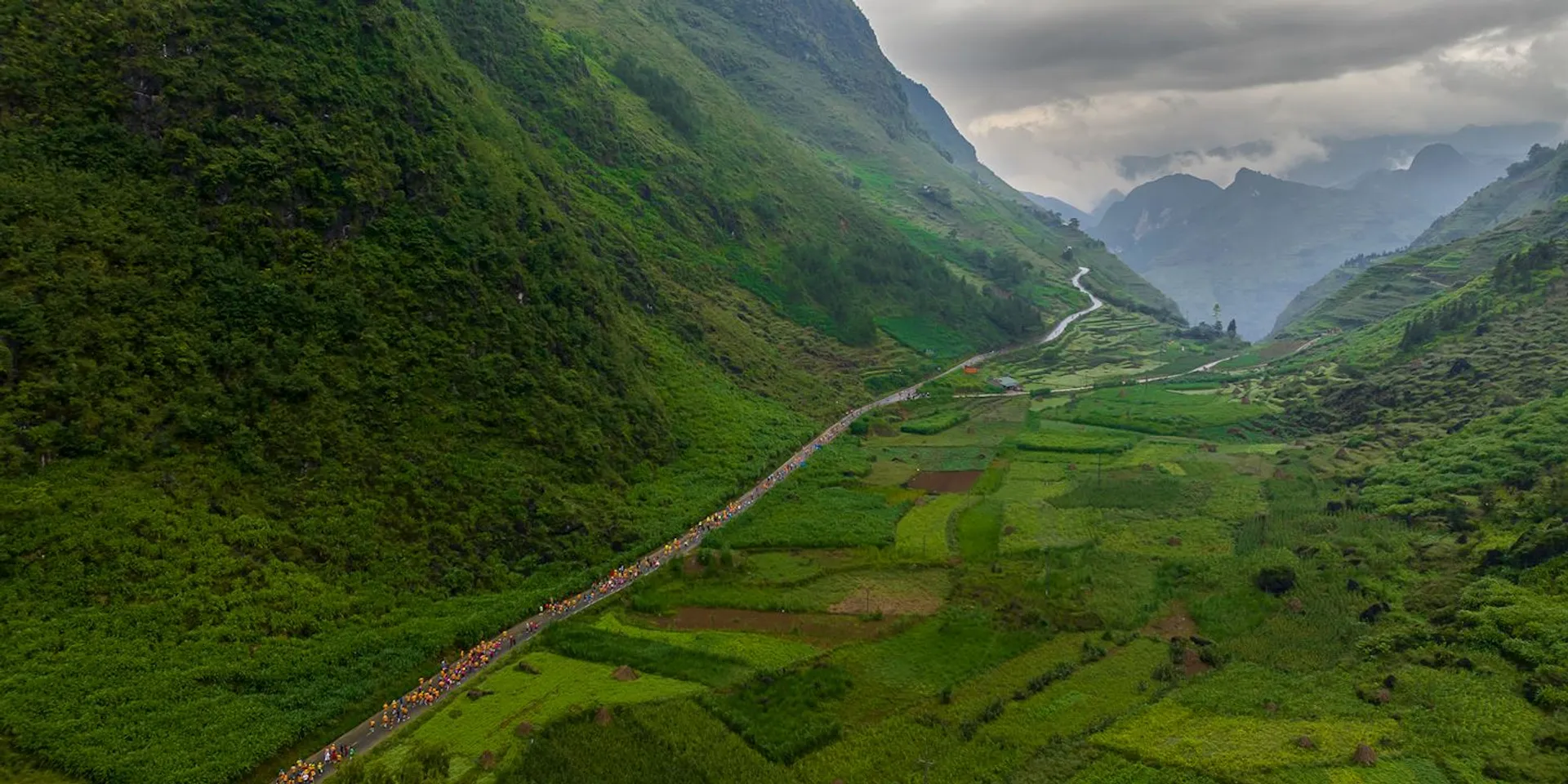 This image shows a lush, green valley with a winding road cutting through it. The valley is bordered by steep, forested hills on both sides. There's a large group of people on the road, likely participating in an event such as a race or a hike. The sky is cloudy, adding a dramatic touch to the landscape.