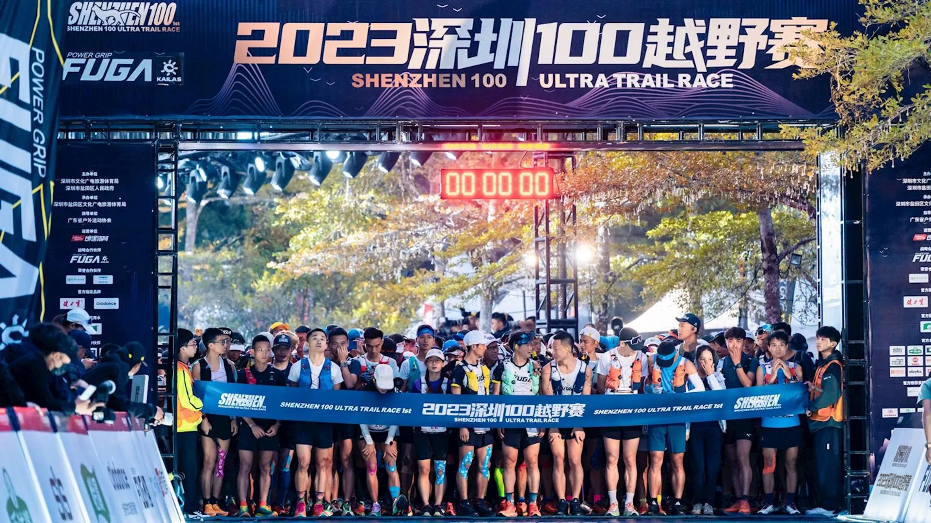 The image shows a gathering of runners at the start line of an event called "Shenzhen 100 Ultra-Trail Race" which occurred in 2023, as indicated by the banner above the starting gate. This event appears to be an ultra-marathon or a long-distance endurance race, as suggested by the "Ultra-Trail" label. Competitors are wearing sportswear typical for runners, including shorts, t-shirts, running shoes, and hydration packs. Many are also wearing caps and sunglasses, suggesting the race may take place in sunny conditions.

A digital clock is displayed at the top center of the starting gate showing "00:00:00," indicating that the race is about to begin or has just started. Banners and signage from