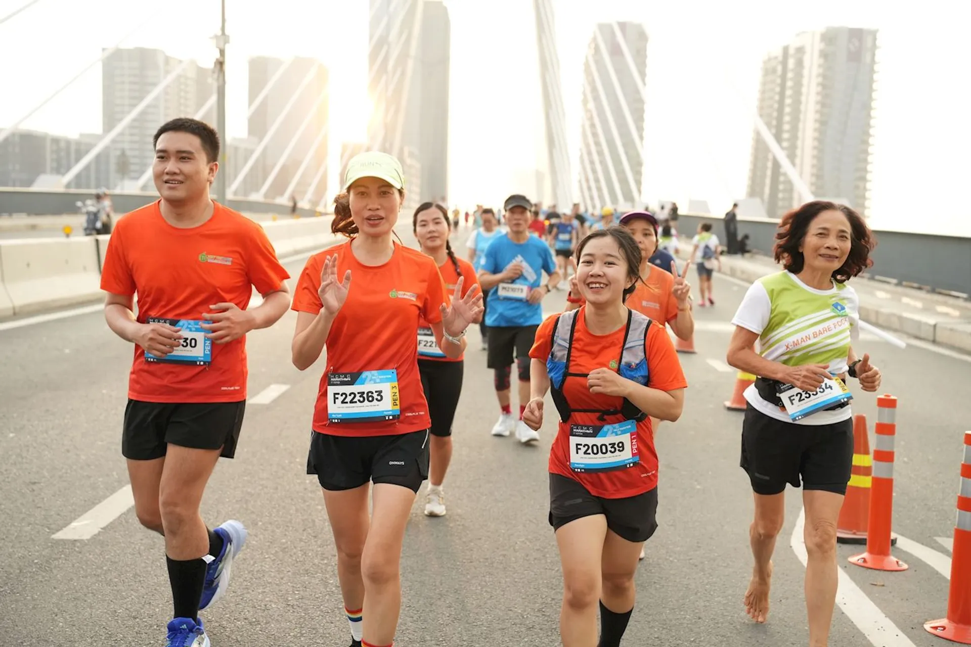 This image shows a group of people running in what appears to be an organized road race or marathon. They are dressed in athletic clothing, with some wearing matching orange shirts and race bibs attached to their fronts, indicating their participation numbers. The runners have various expressions, some smiling, suggesting they are enjoying the event despite the physical challenge. The background features a modern bridge with distinct architecture, and there is a clear sky which might indicate good weather for the event. It seems to be a lively and well-attended occasion focused on health, fitness, and community involvement.
