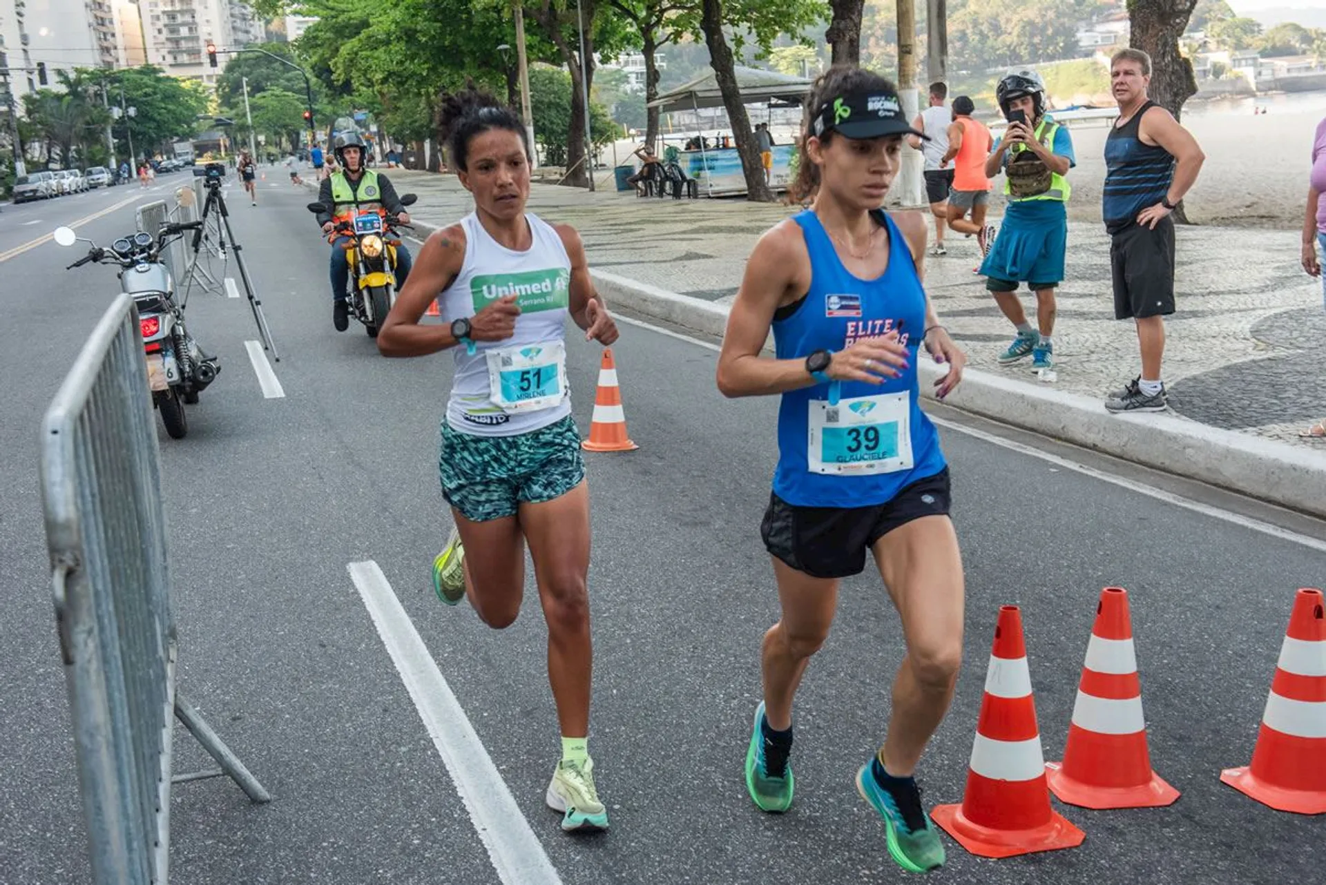 In this image, we see two female athletes participating in a running event, each wearing a bib indicating their participant numbers (51 and 39). They are in motion, suggesting they are in the middle of the race. The setting appears to be an urban area with trees and buildings in the background, and the street is partially closed for the race with orange traffic cones. There are onlookers and other participants in the background, as well as a motorcycle, indicating that there might be race officials monitoring the event. The runners are attired in sports gear appropriate for a competition—running shoes, shorts, and tank tops—with one wearing a visor cap. The focus and determination on their faces suggest they are pushing towards a strong finish.