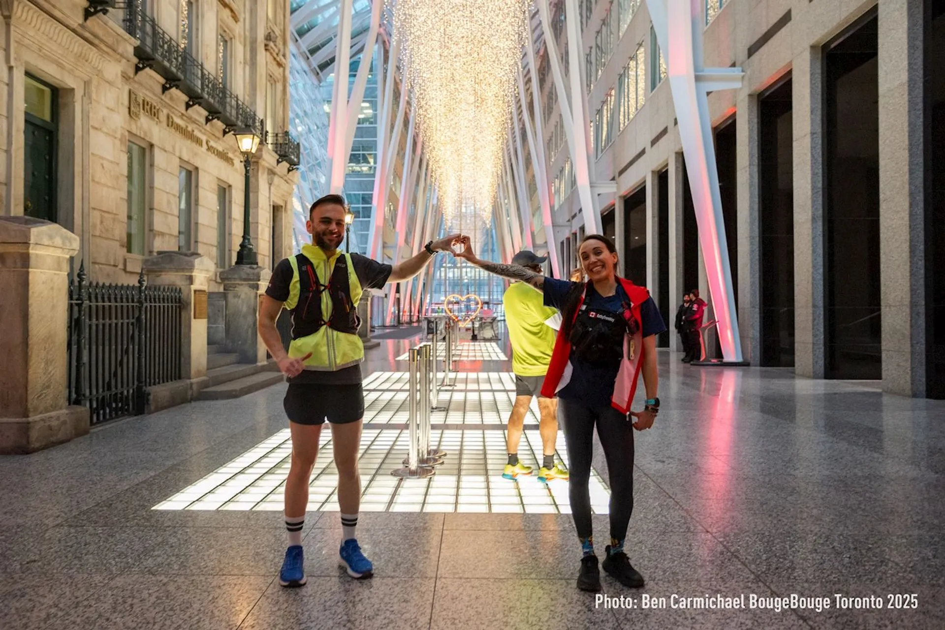 The image shows two people, a man and a woman, standing on a brightly lit path inside a building with glass walls and a high ceiling. They are facing the camera and holding hands, smiling. Both are wearing athletic clothing and have backpacks, suggesting they might be involved in a running or fitness activity. The setting appears to be a modern, architecturally striking indoor space.
