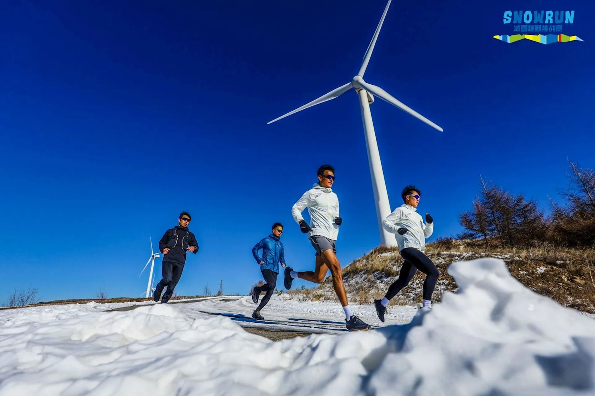 This image depicts a group of runners (four in view) jogging through a snowy landscape with a clear blue sky overhead. They appear to be on a trail with patches of snow and grass visible, indicating that the snow is not very deep, or perhaps it is melting. In the background, there is a large wind turbine, indicative of a wind farm or a location where renewable energy is harnessed. The runners are dressed in athletic gear suitable for cold weather, with long pants and jackets. The presence of the "Snowrun" watermark suggests that this might be part of an organized running event or race taking place in a wintry environment.